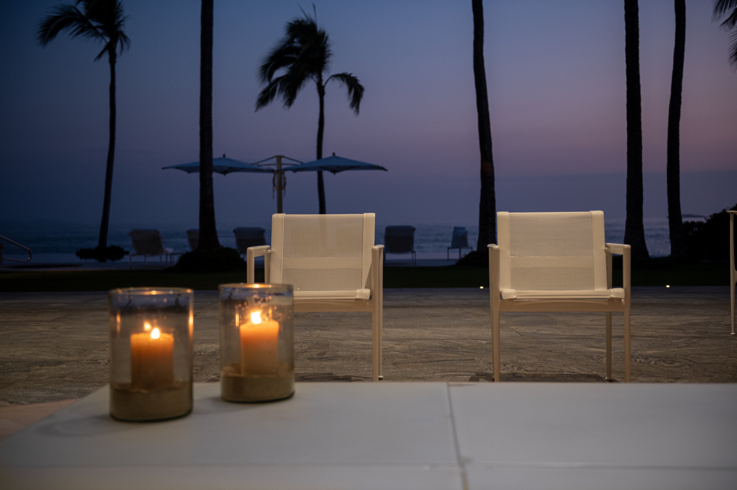 A beachside setting at dusk with tall palm trees silhouetted against a purple and blue sky, outdoor chairs, and lit candles on a table in the foreground.