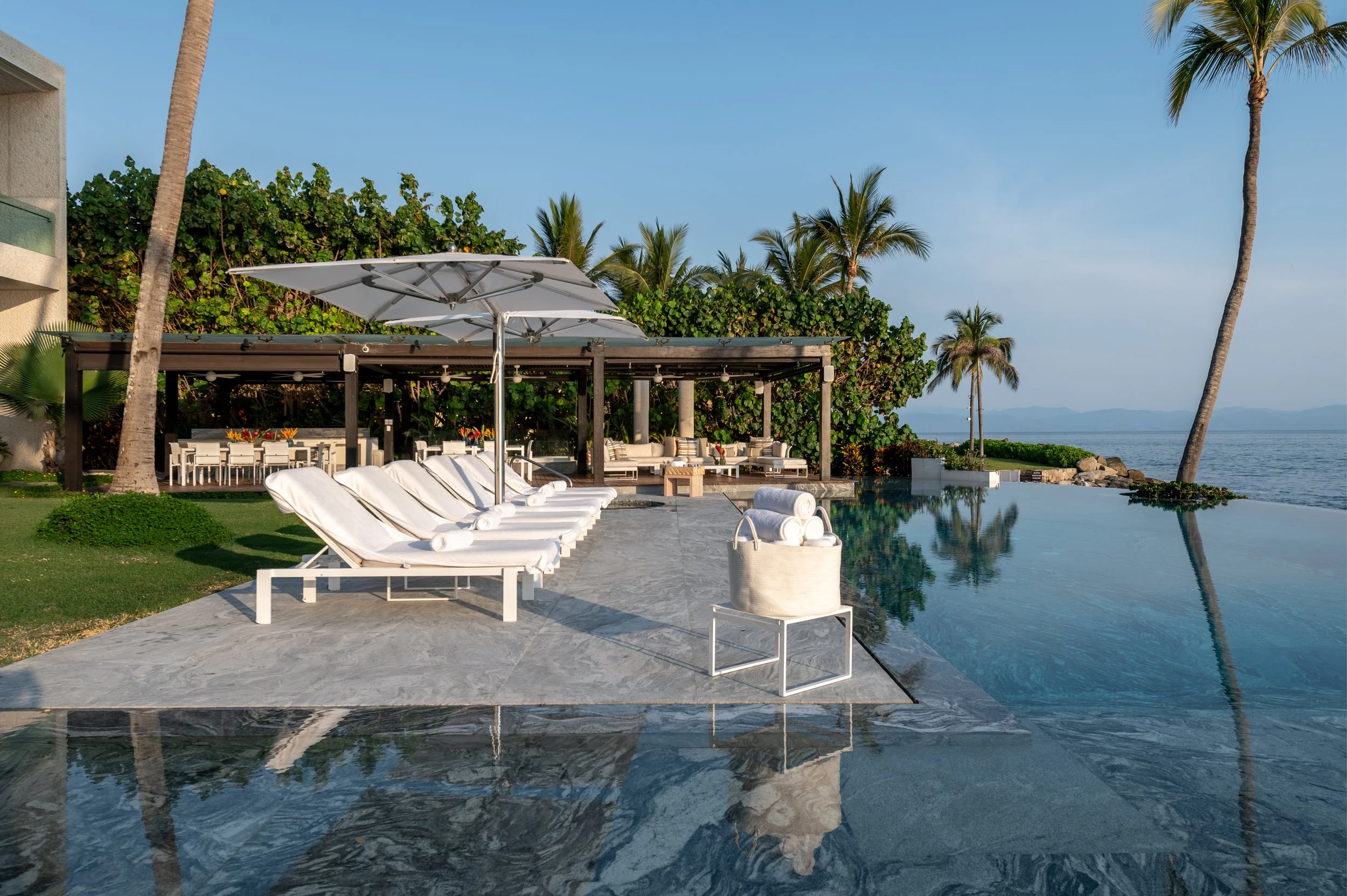 A luxurious outdoor pool area with white lounge chairs, large white umbrellas, and patio furniture under a shaded pergola, overlooking the ocean with palm trees and a distant island in the background.