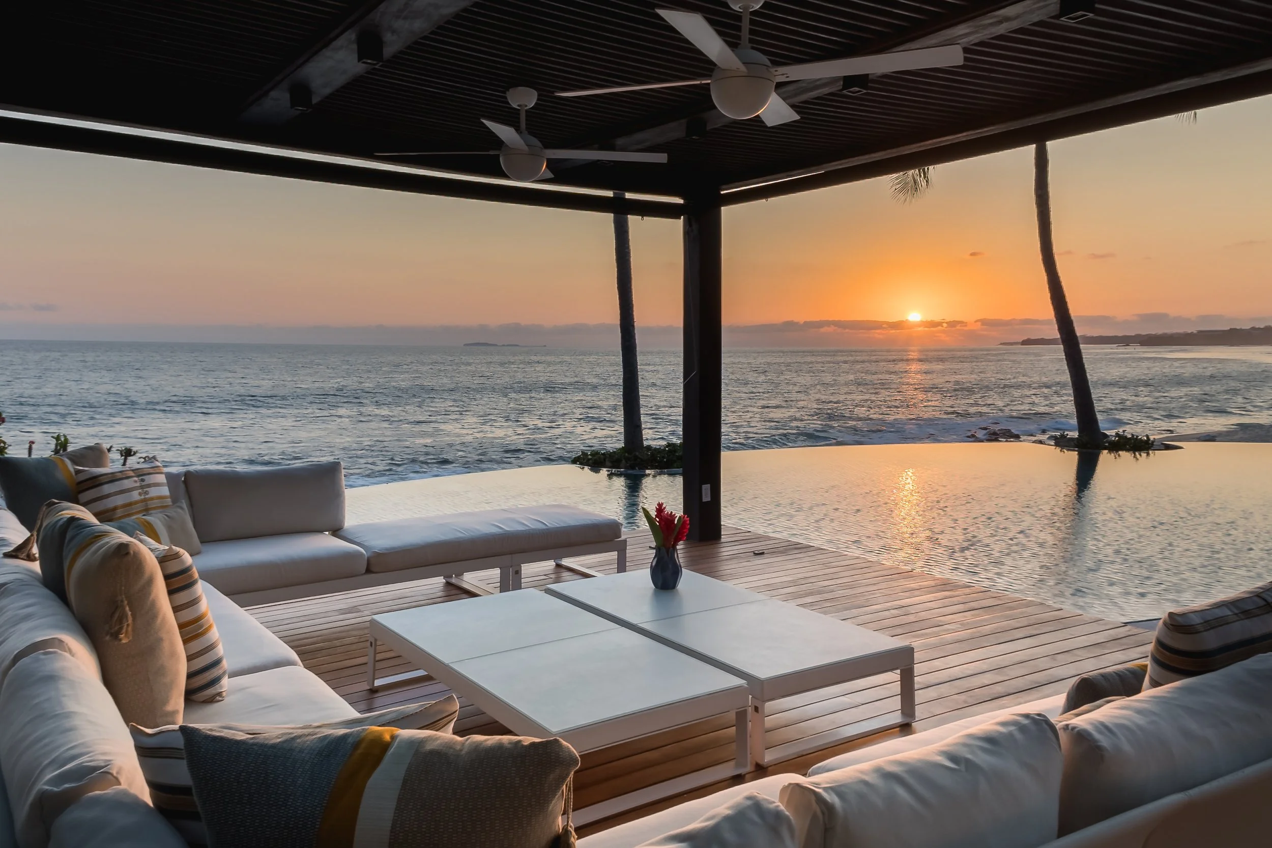 Outdoor seating area with a white sofa and pillows, overlooking the ocean during sunset, with a ceiling fan and a view of the horizon.