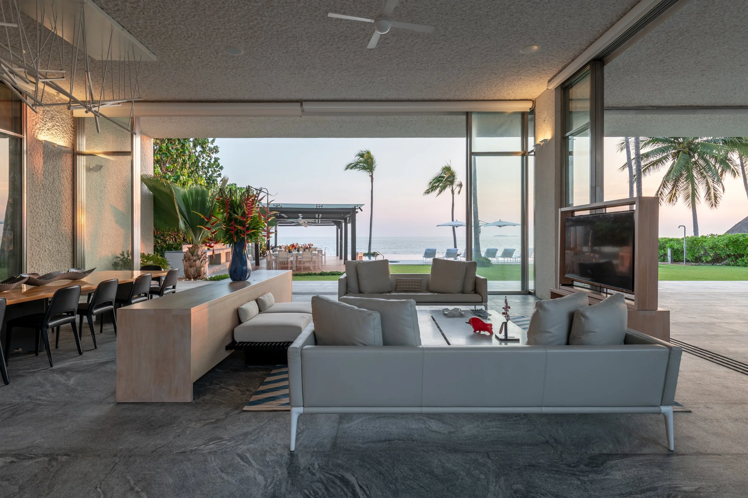Living room with large glass sliding doors opening to a beachside view, featuring outdoor lounge chairs, palm trees, and a sunset sky.