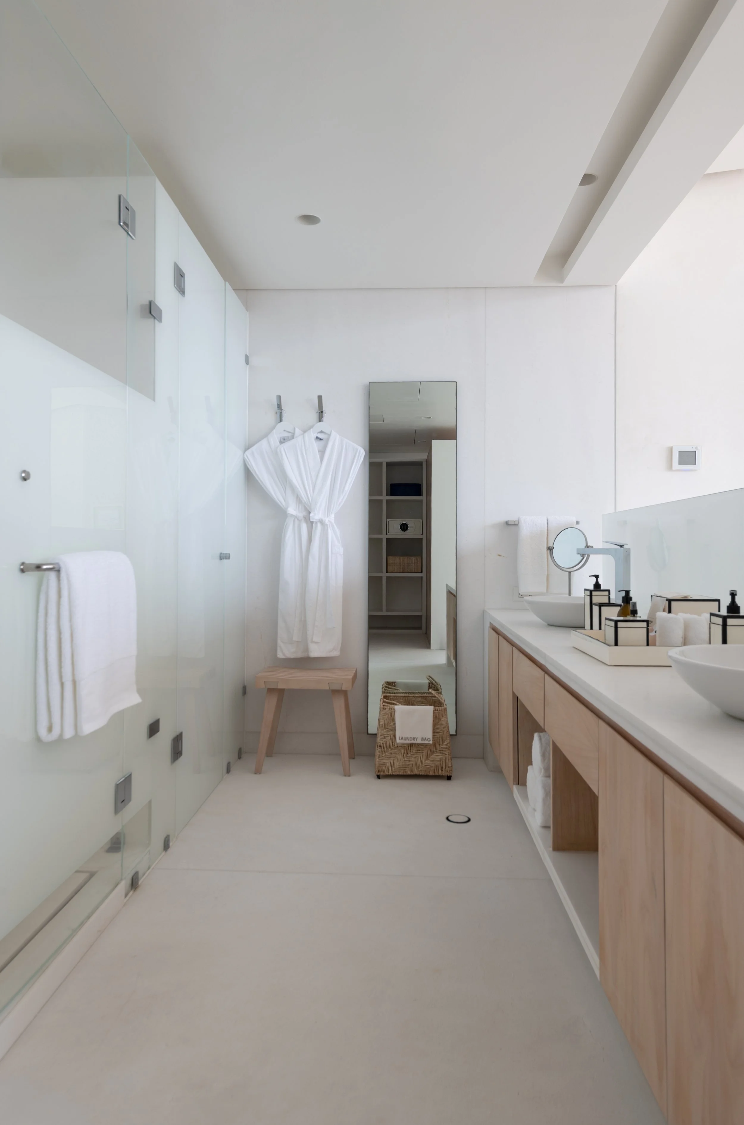 Modern bathroom with white walls, a large mirror, a wooden vanity with a vessel sink, and various toiletries. White towels hang on hooks, and a robe is on a stool. A laundry basket sits on the floor, and a small mirror is on the countertop.