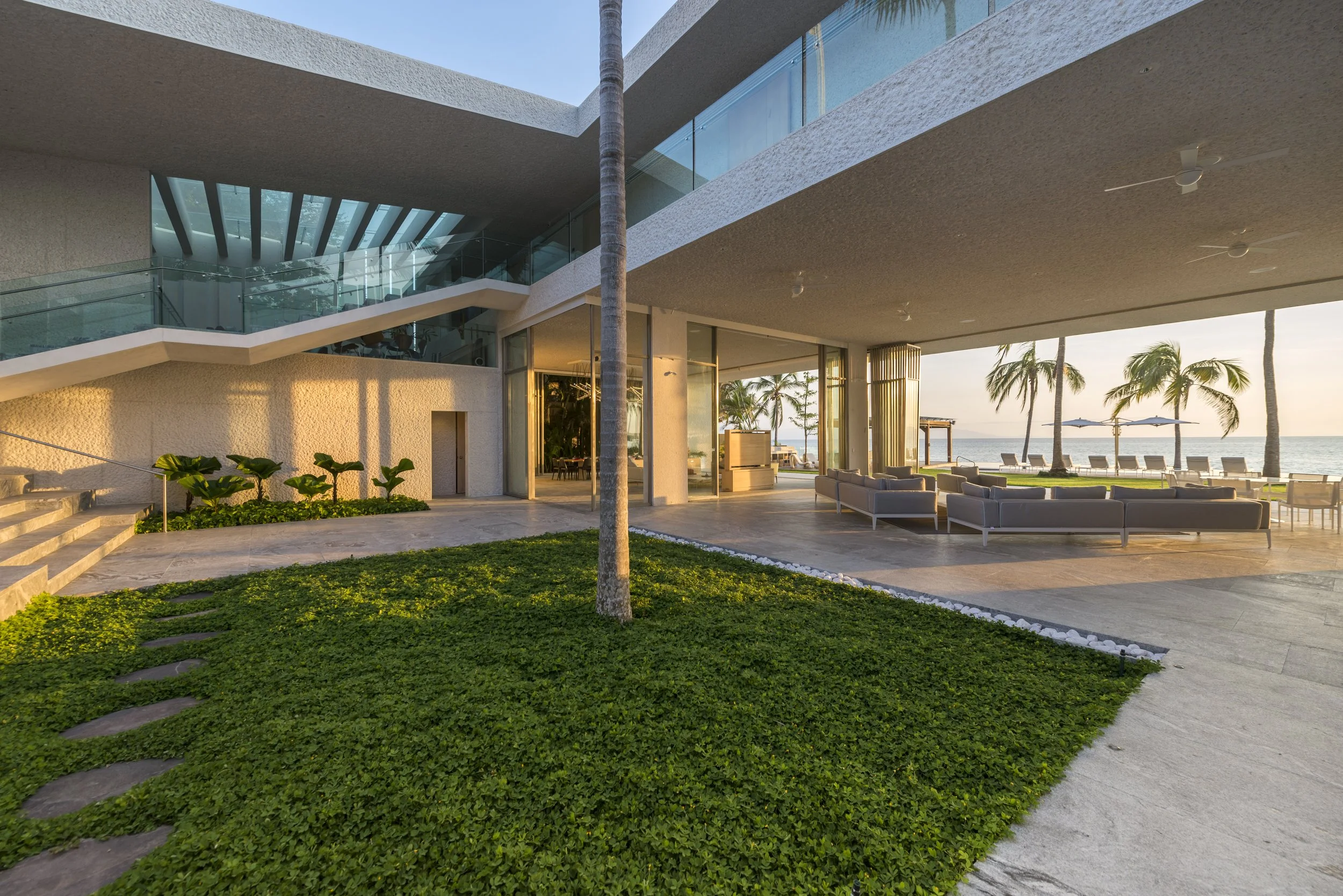 Modern beachfront building with outdoor seating area, palm trees, and ocean view at sunset.