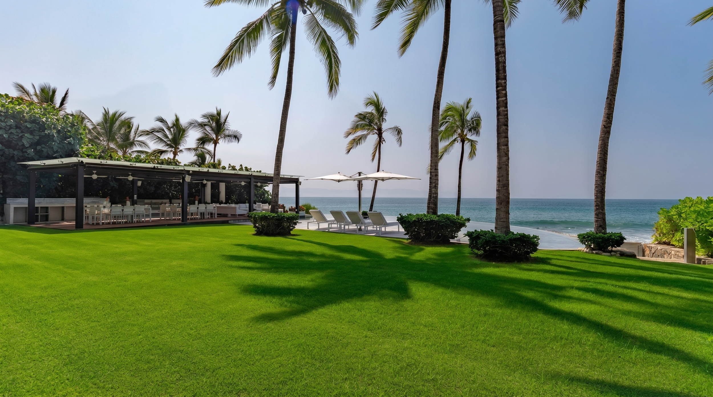 A tropical beachfront scene with palm trees, green lawn, lounge chairs with umbrellas, and a view of the ocean.