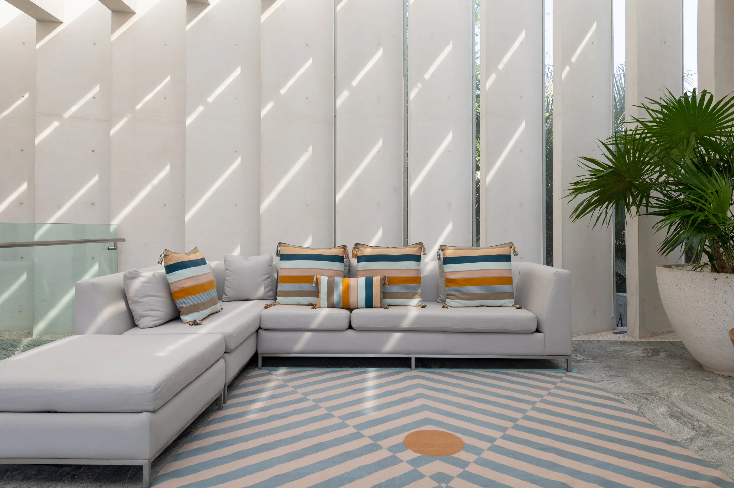 Modern indoor lounge with white sectional sofa and striped pillows, large windows with sunlight patterns, green potted plant, geometric patterned rug.