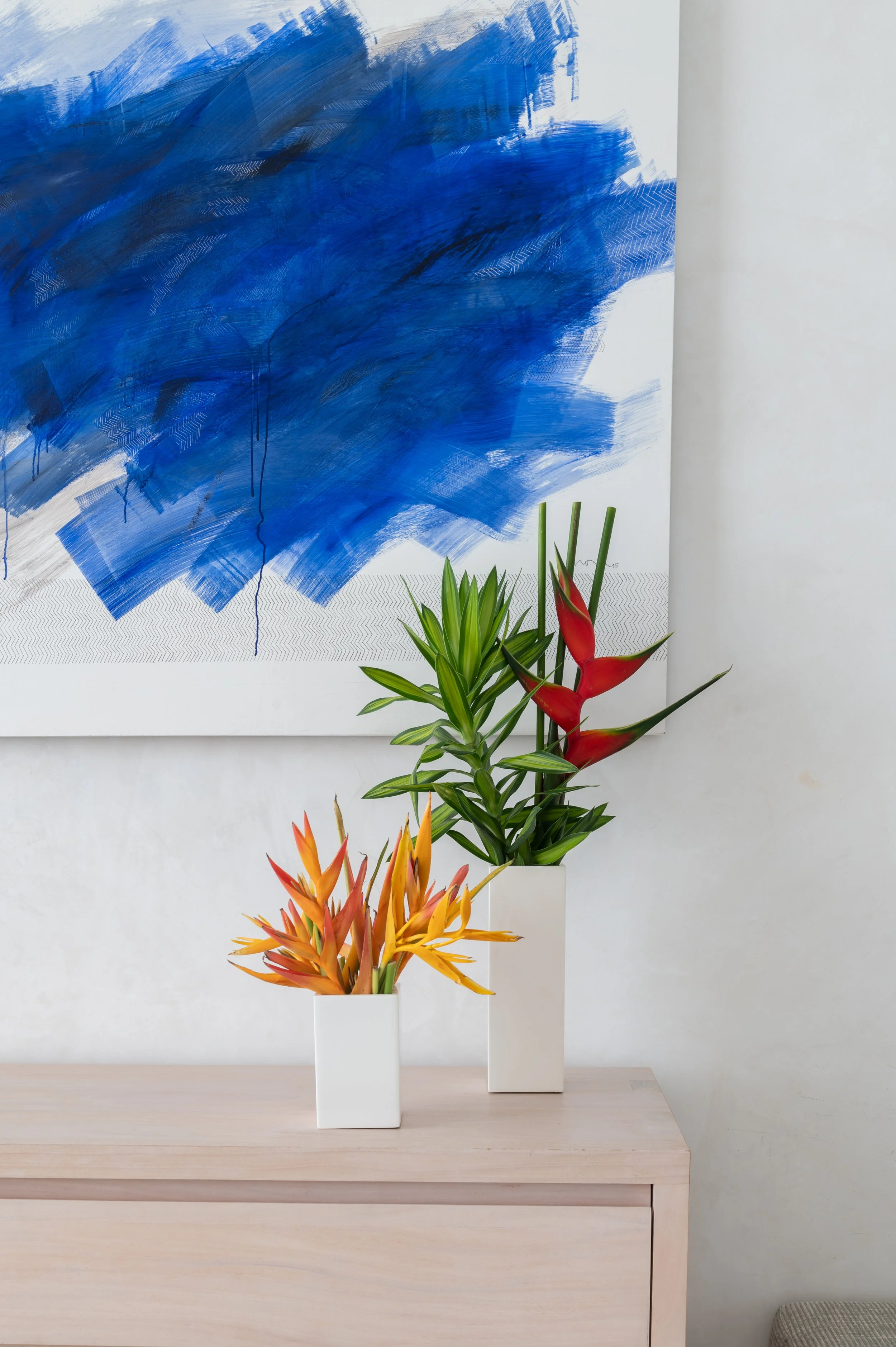 A modern, minimalist room with a light wooden table, two white vases with tropical flowers, an abstract blue painting on the white wall, and a light gray cushion on the floor.