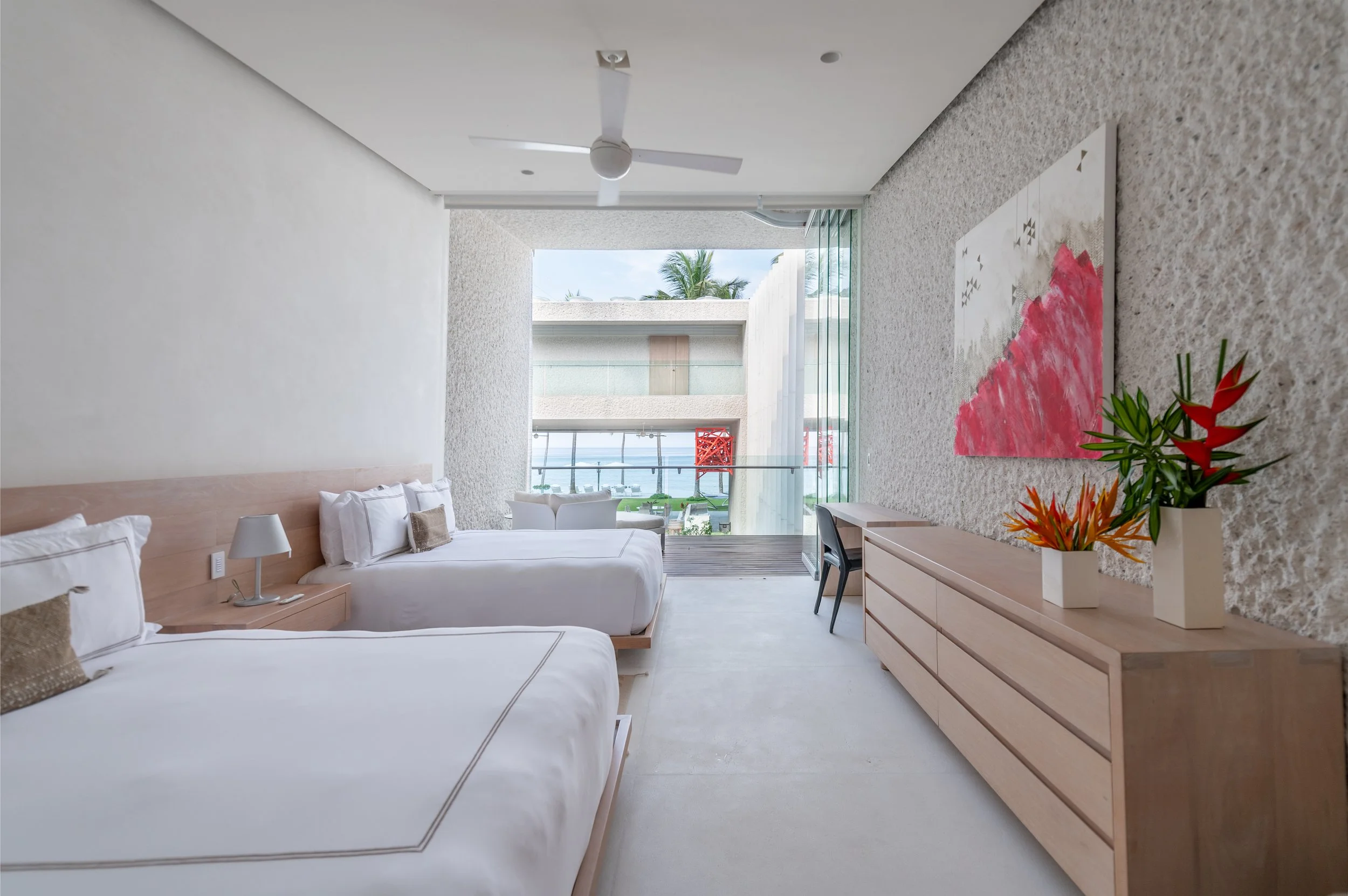 A modern hotel room with two white beds, a nightstand with a lamp, a wooden dresser with vases of colorful flowers, a desk with a chair, and a large window revealing an outdoor balcony and a view of palm trees and blue sky.