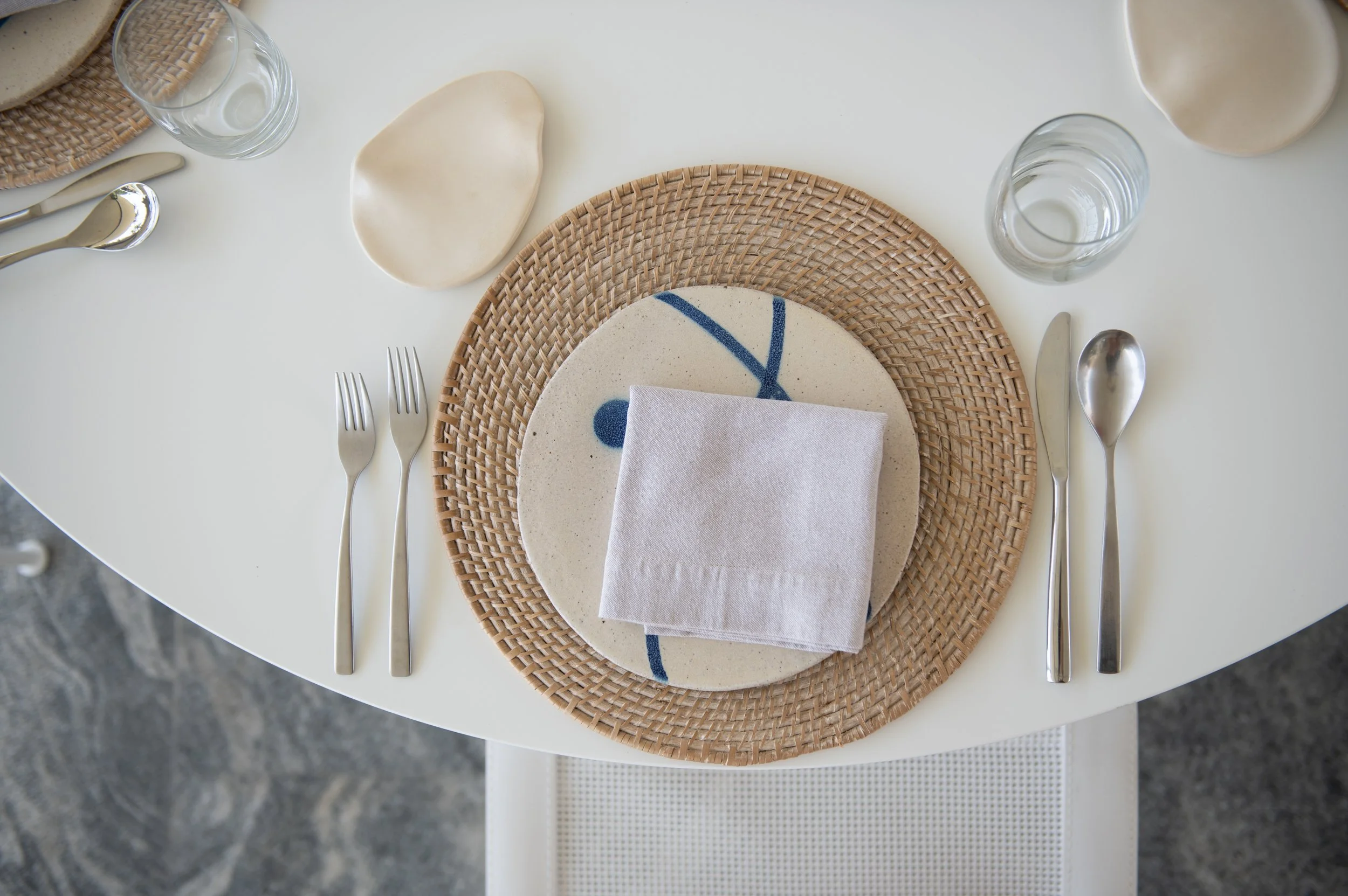 A minimalist table setting with a round white table, a woven placemat, a decorative ceramic plate with blue stripe design, beige cloth napkin, and silverware. There are also two glasses of water and a ceramic bowl or jug, with a wavy-edged clay or ce