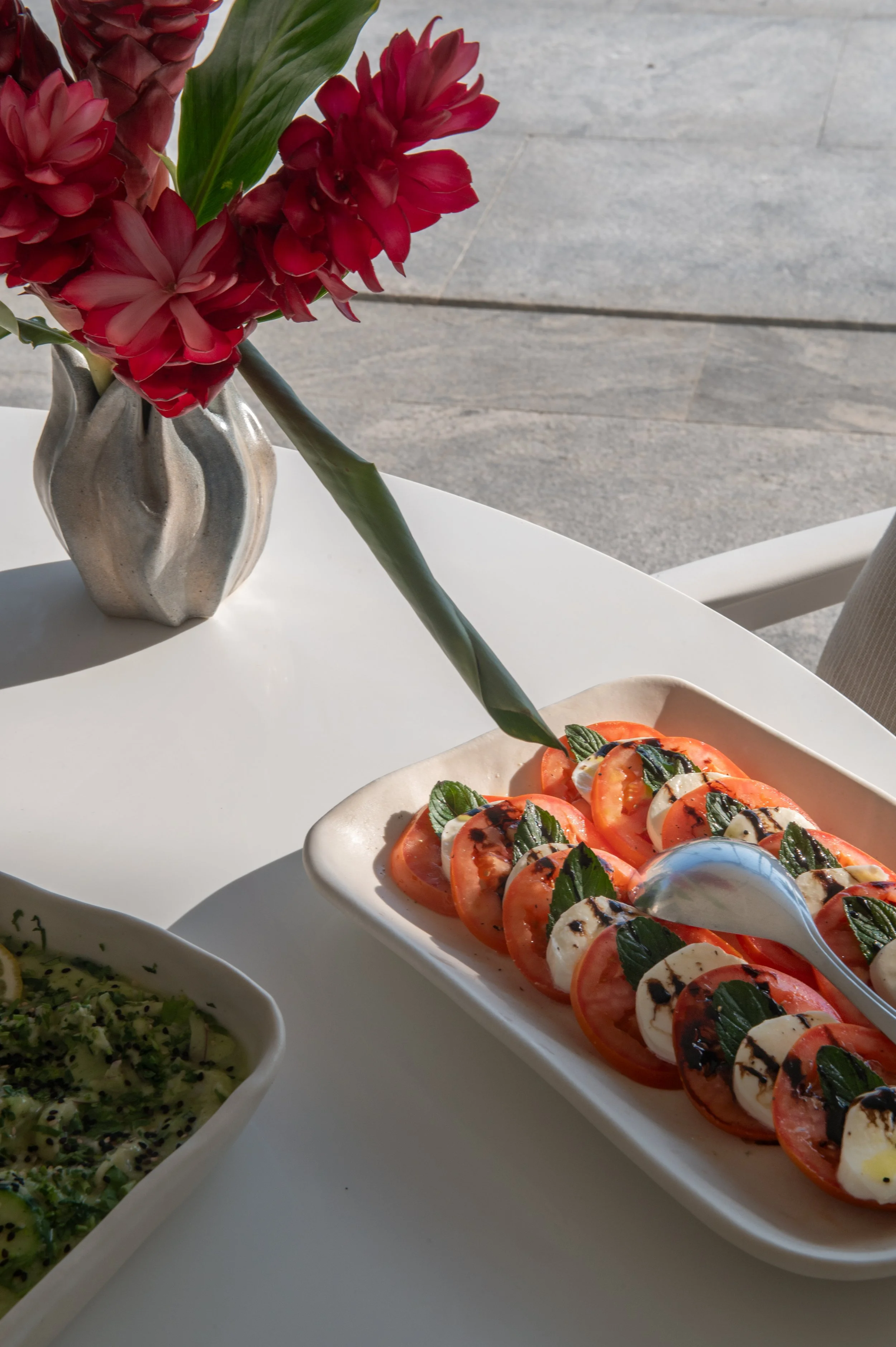 Close-up of a plate with sliced tomatoes, mozzarella, basil, and balsamic glaze, on a white table, with a vase of red flowers in the background.