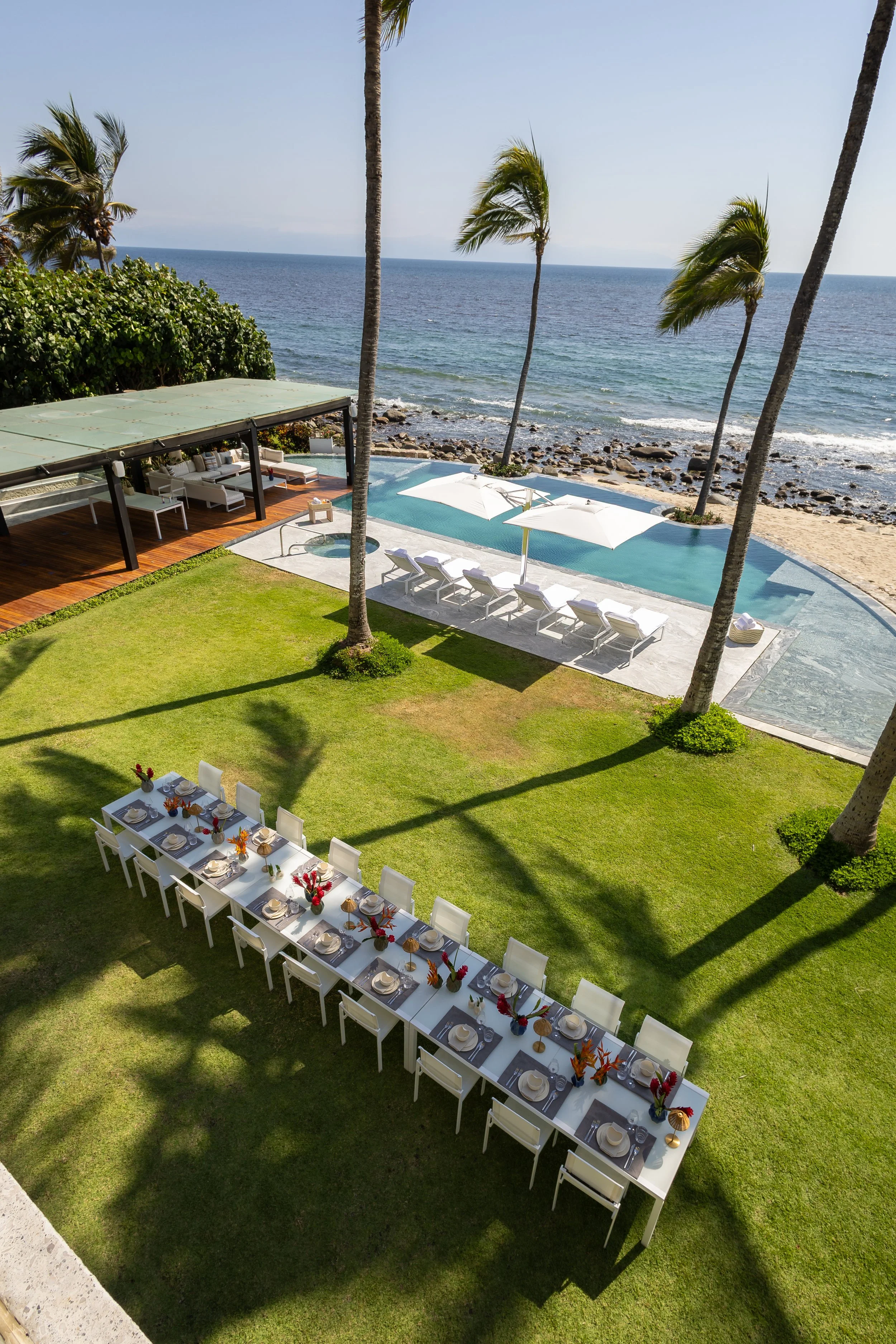 Beachside outdoor dining setup with a long white table decorated with flowers, set for a meal, overlooking a lush green lawn, pool, rocky shoreline, ocean, and palm trees under a clear sky.