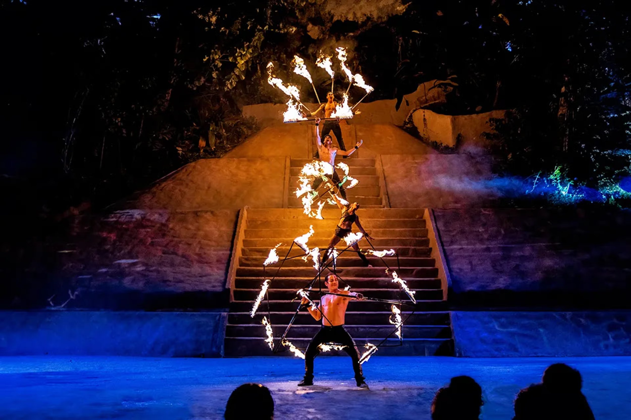 A nighttime performance featuring a group of fire performers on steps, with staff and poi flaming, surrounded by dark trees and colorful lighting effects.
