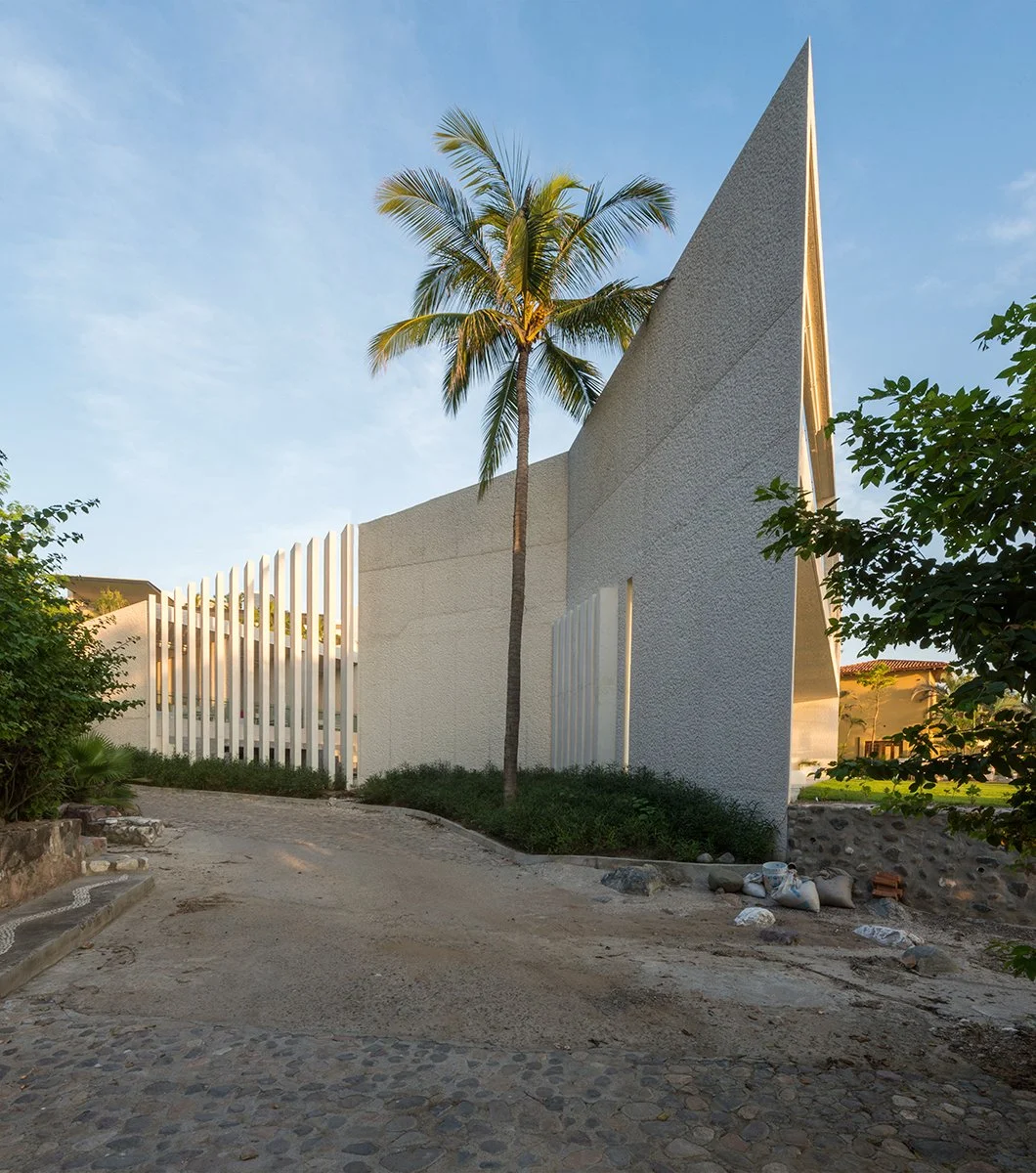 Modern building with geometric design and textured white walls, surrounded by tropical greenery, including a tall palm tree, under a clear blue sky.