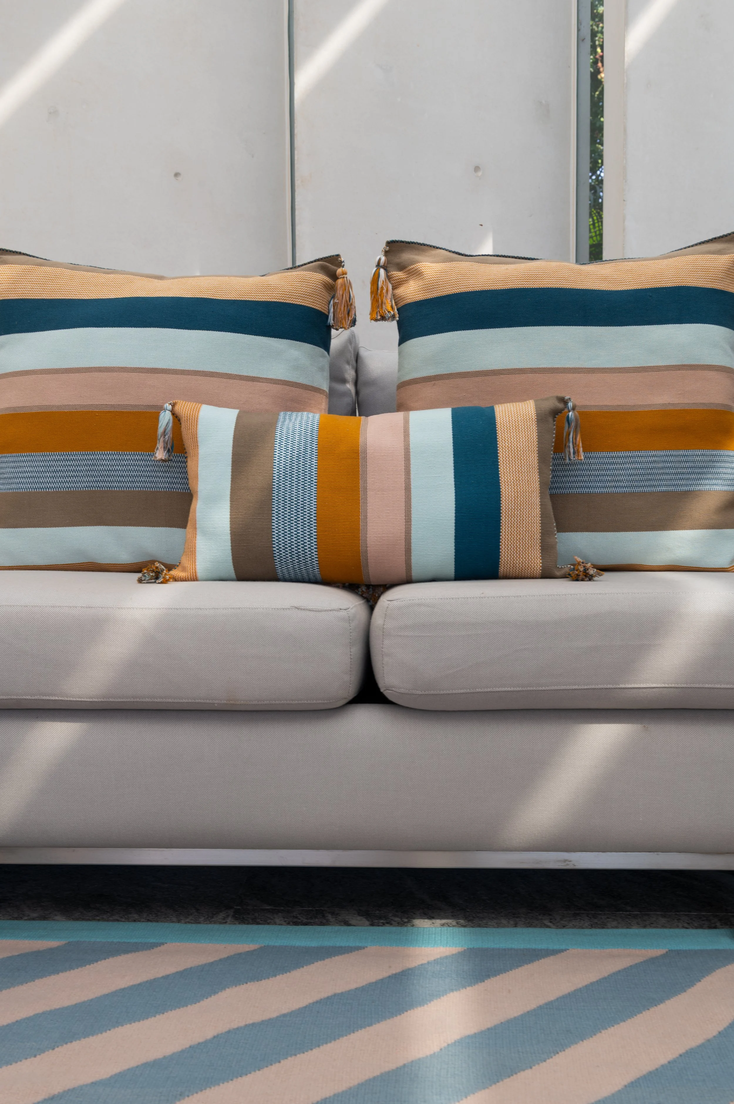 Two striped pillows with tassels on a sofa in front of a white wall and a window with plants outside, part of a sitting area with a gray sofa and patterned rug.