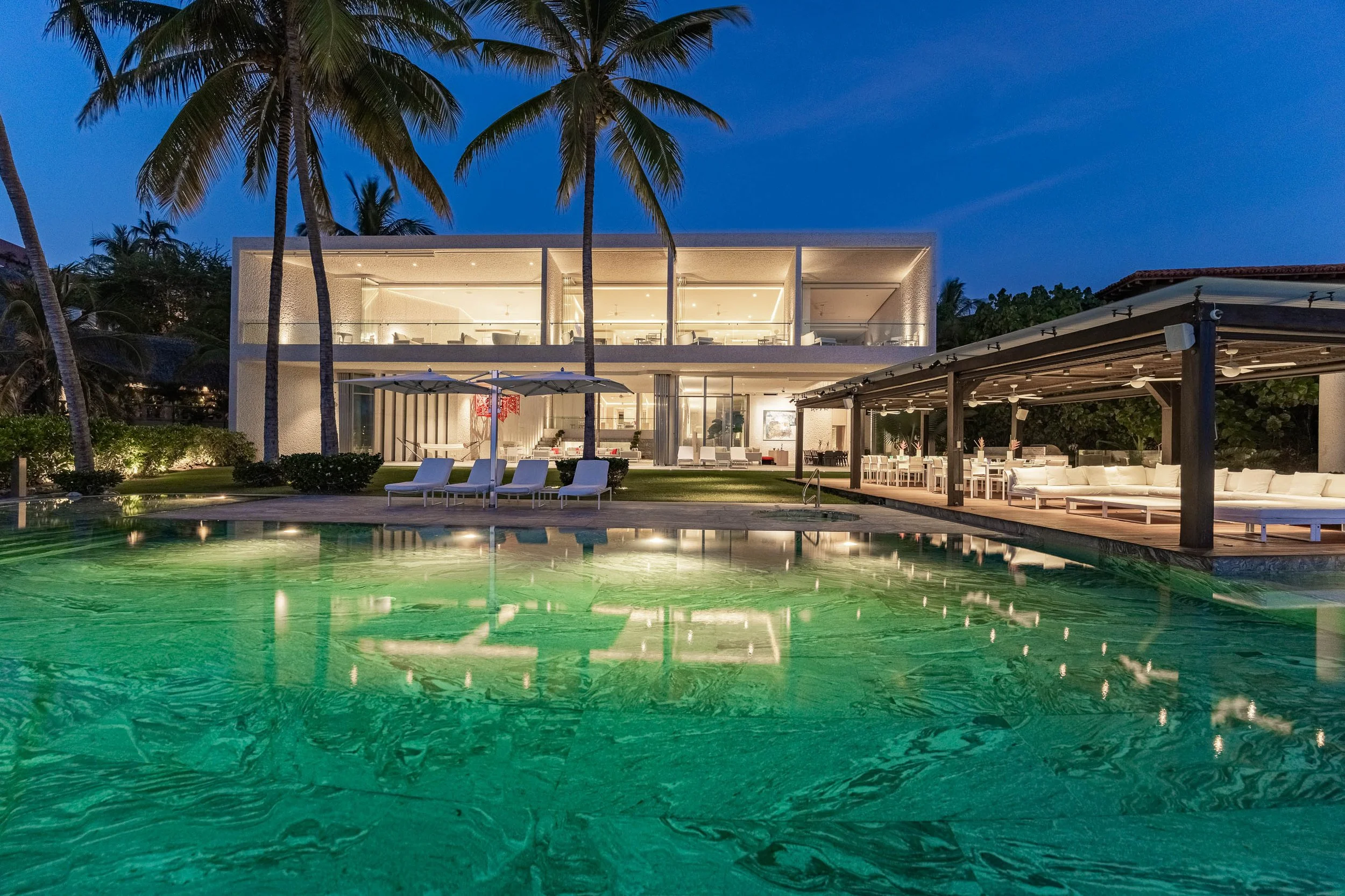 Modern house by a lit pool with palm trees and outdoor sitting areas at night.