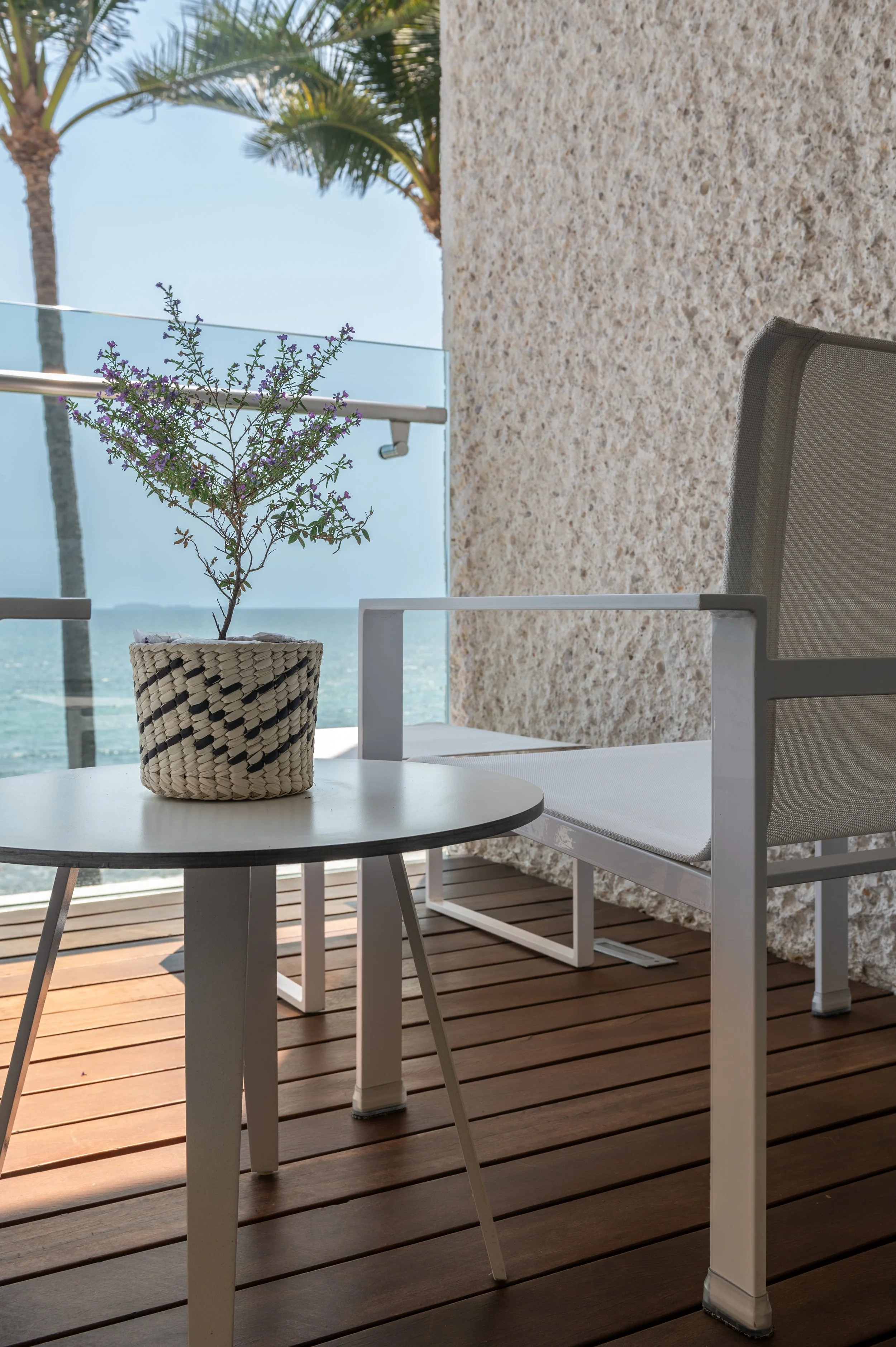 A white table with a potted purple flowering plant, a white chair, a glass railing, palm trees, and a view of the ocean.