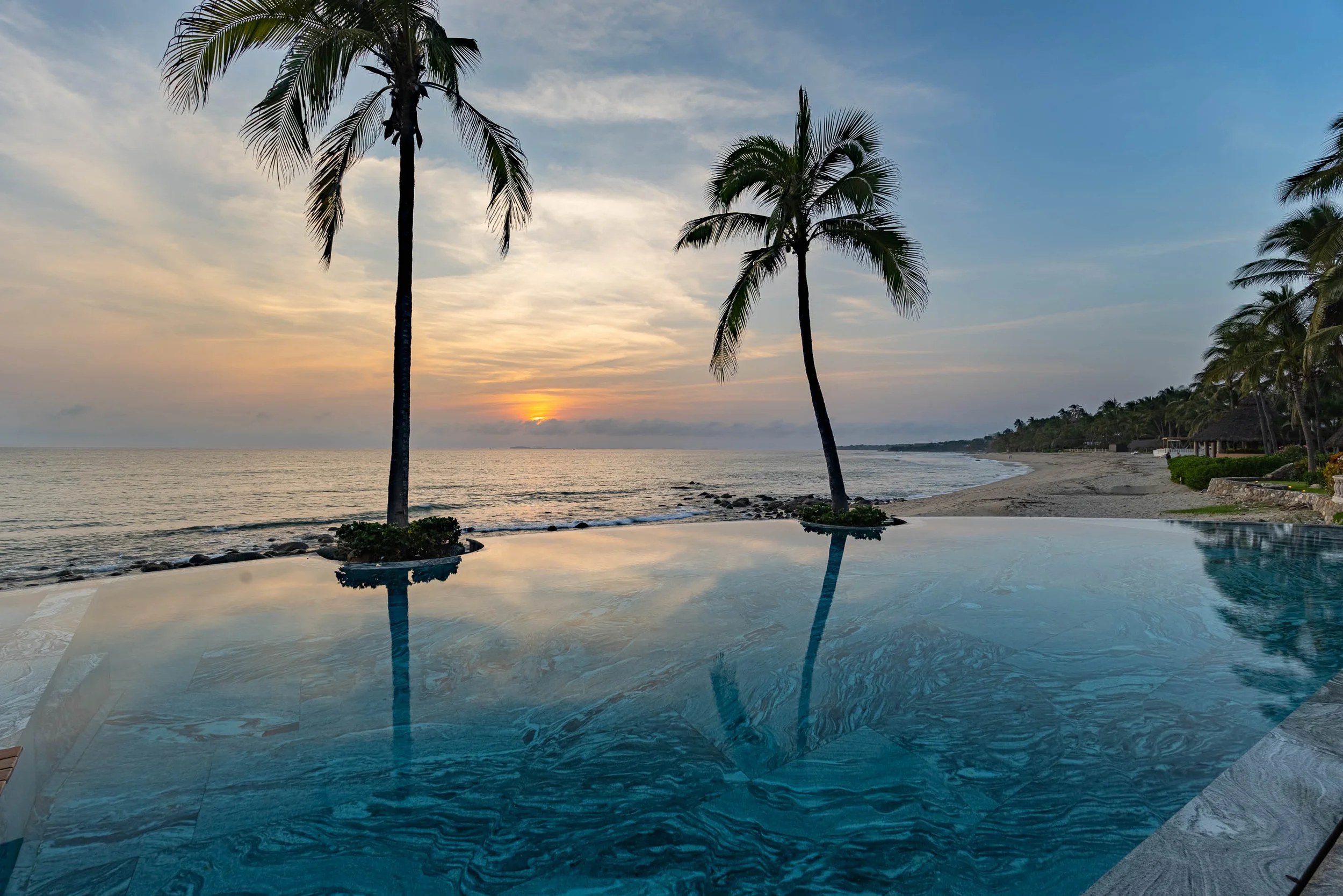 A sunset over the ocean viewed from a beach, with tall palm trees in the foreground and a reflective swimming pool in the bottom part of the image.