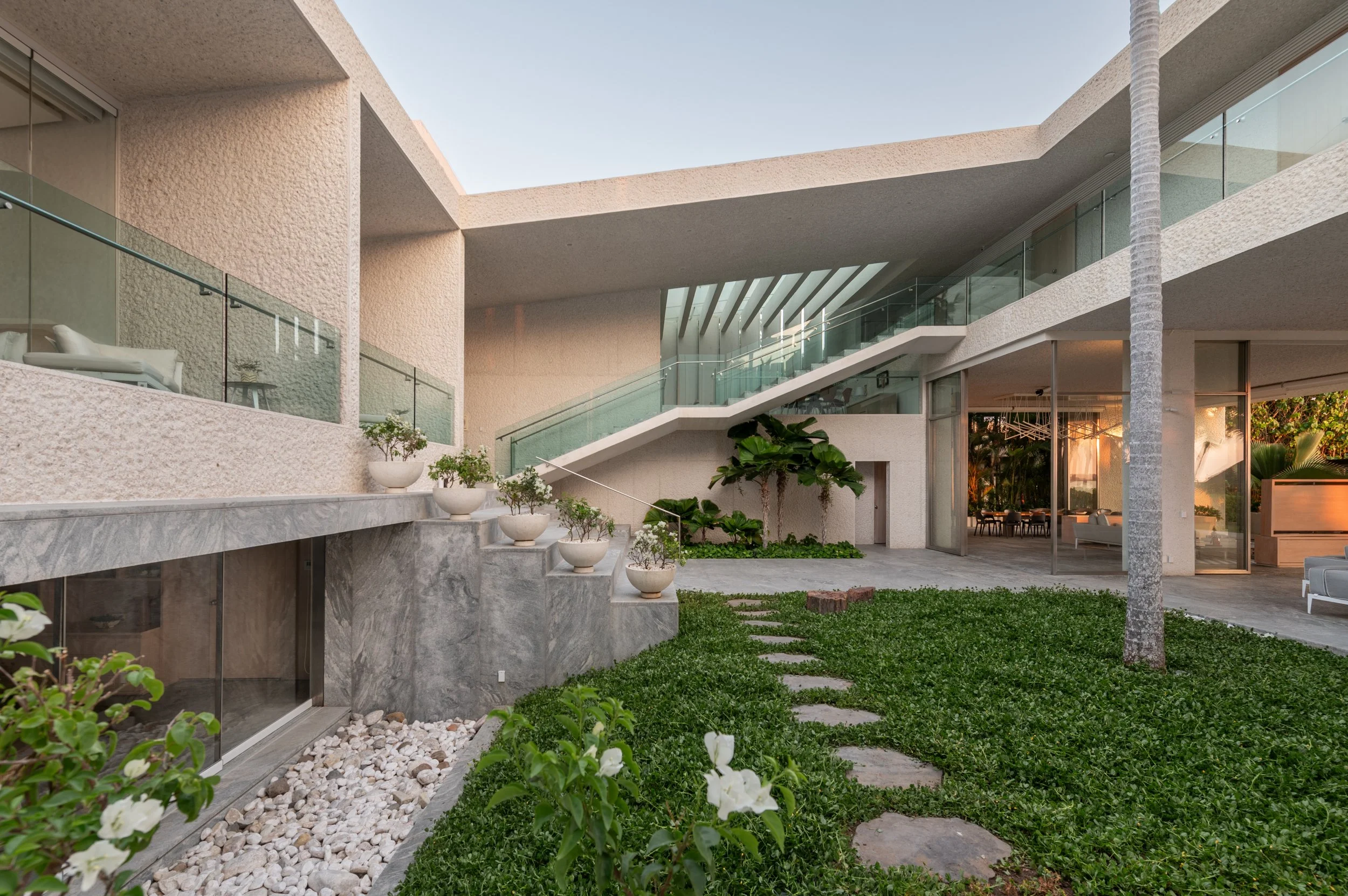 Modern house with outdoor patio, glass railing, staircase, potted plants, and a grassy yard in the evening.