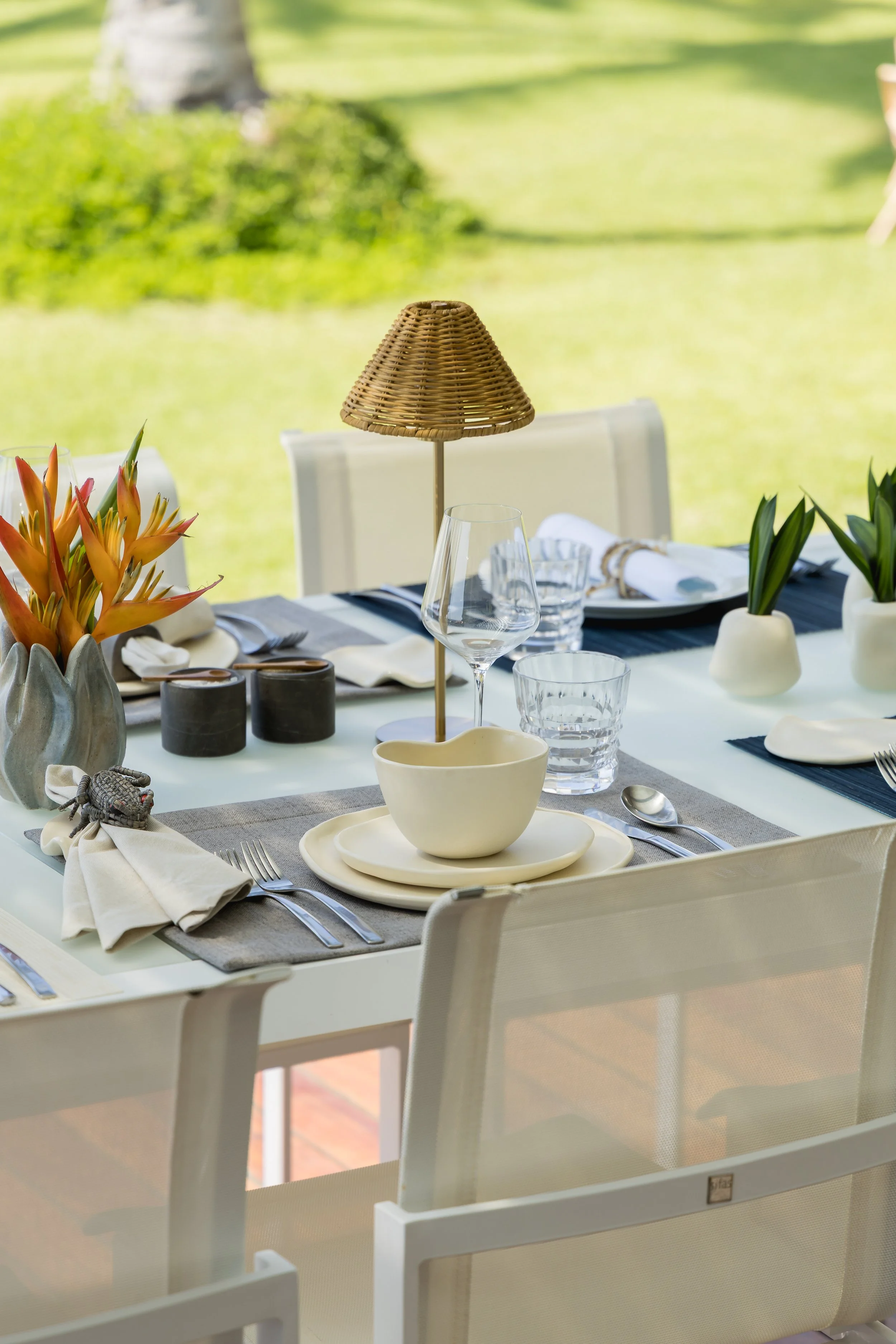 Dining table set with white china, crystal glassware, silverware, napkins, and decorative plants, with a rattan table lamp in an outdoor setting with lush greenery.