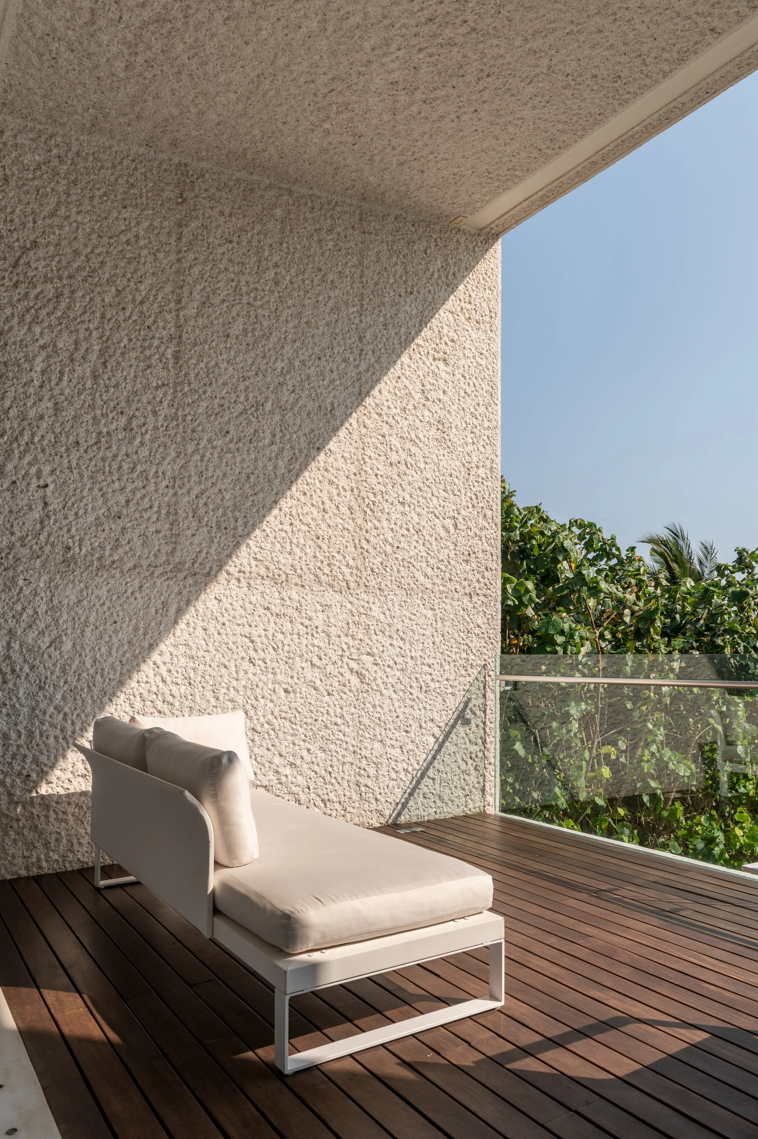 A white outdoor lounge chair with a cushion, situated on a wooden deck next to a textured beige wall, with lush green trees and a clear blue sky in the background.