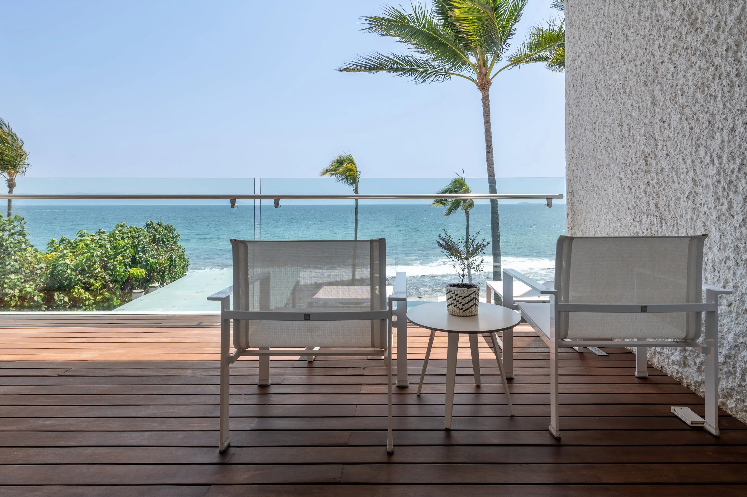 View from a balcony with white outdoor chairs and a small round table with a potted plant, overlooking the ocean with palm trees and a clear blue sky.