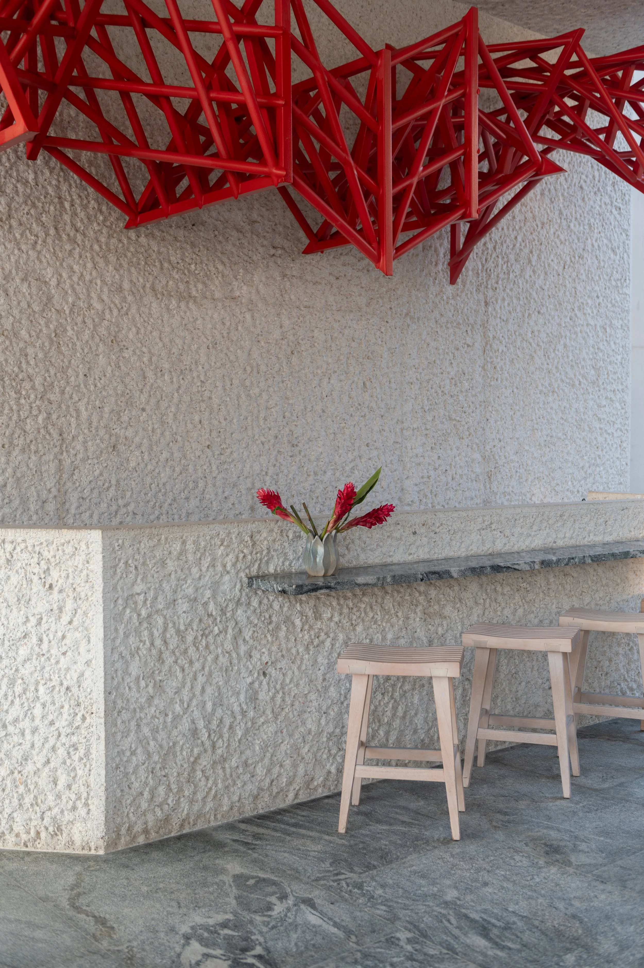 A textured beige wall with a red decorative wall sculpture made of interconnected triangular metal pieces. In front of the wall, there is a small white vase with pink flowers placed on a gray stone ledge. Two light wooden stools are positioned beside