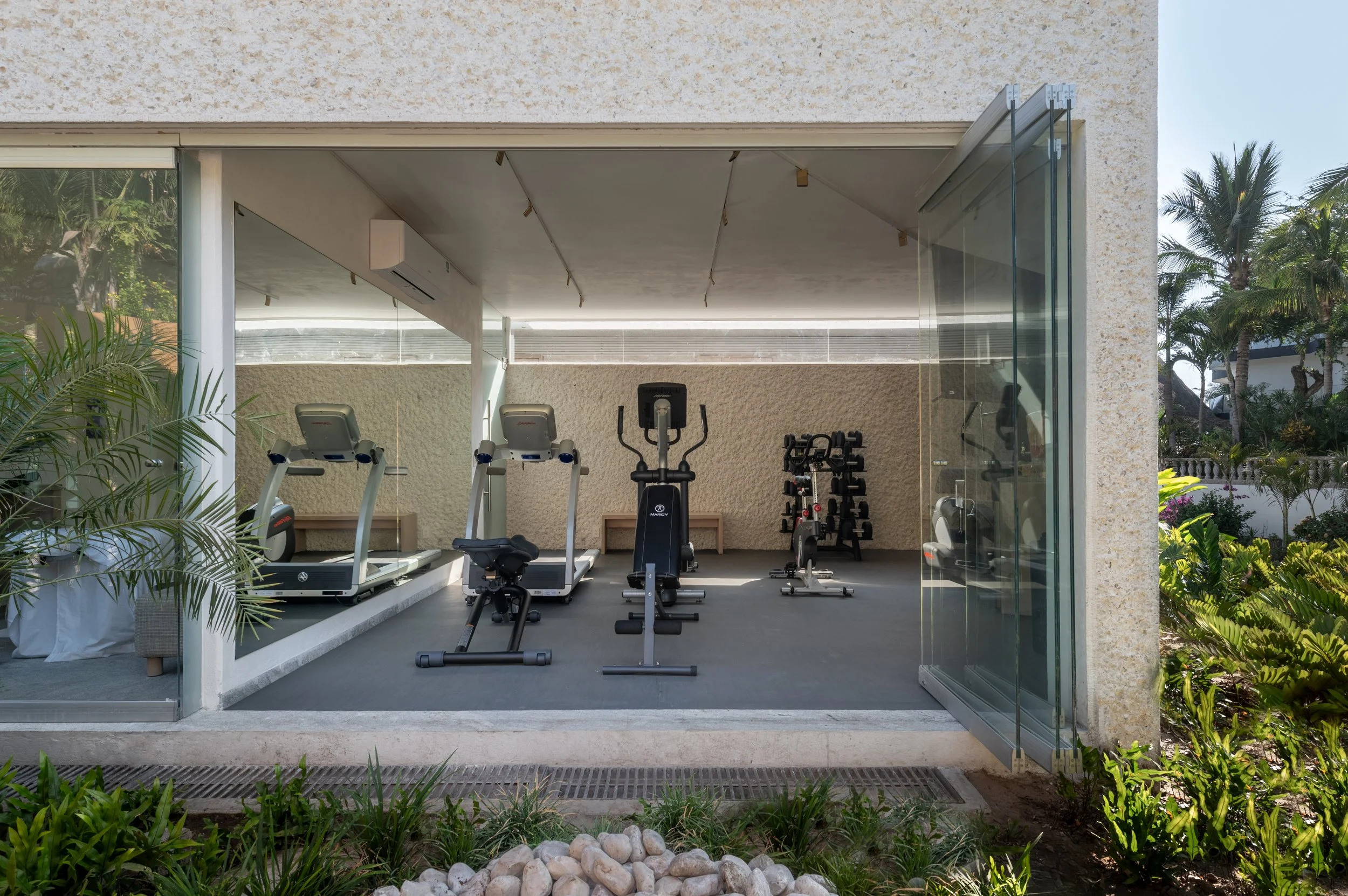 An indoor gym with exercise equipment including treadmills, ellipticals, a stationary bike, dumbbells, and workout benches, seen through an open glass wall with greenery outside.