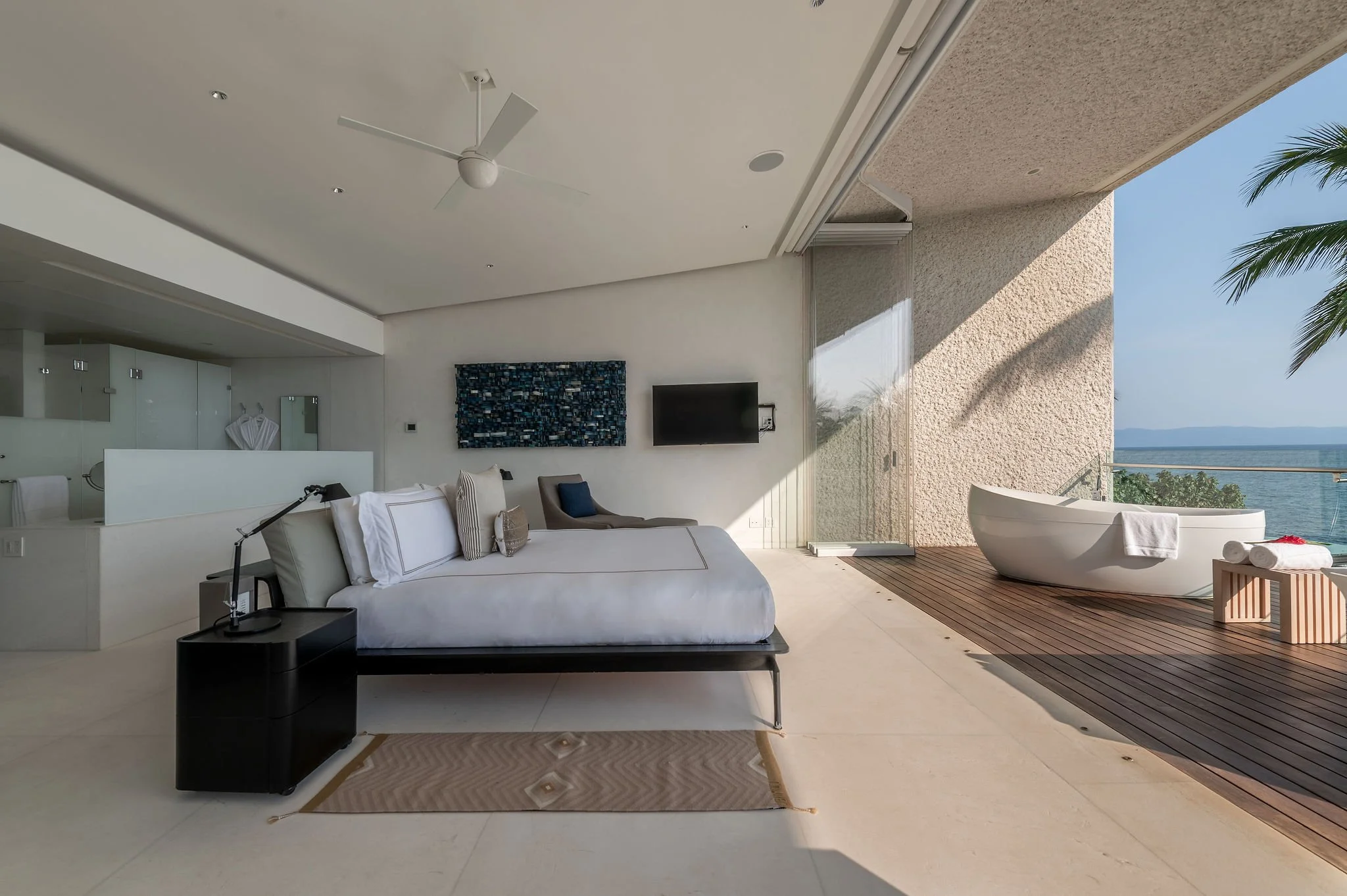 Modern living room with white sofa, black coffee table, wall-mounted television, artwork, and a sliding glass door opening to a balcony with ocean views and a backdrop of blue sky and palm trees.
