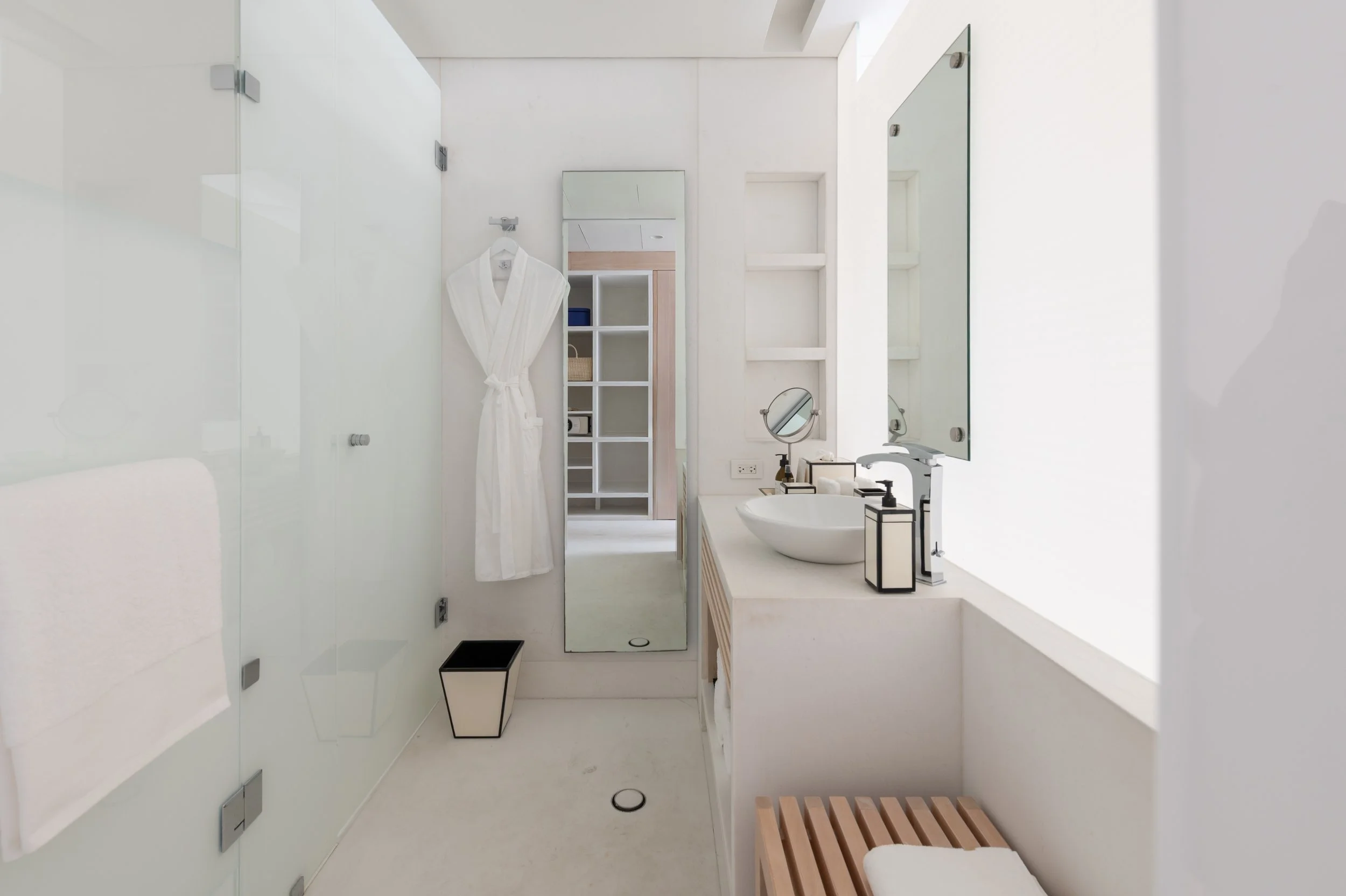A modern, minimalist bathroom with white walls, a white countertop with a round vessel sink, a large mirror, and a white bathrobe hanging on a hook. There are small black accent pieces and a wooden bench with a white towel at the foreground. A tall w