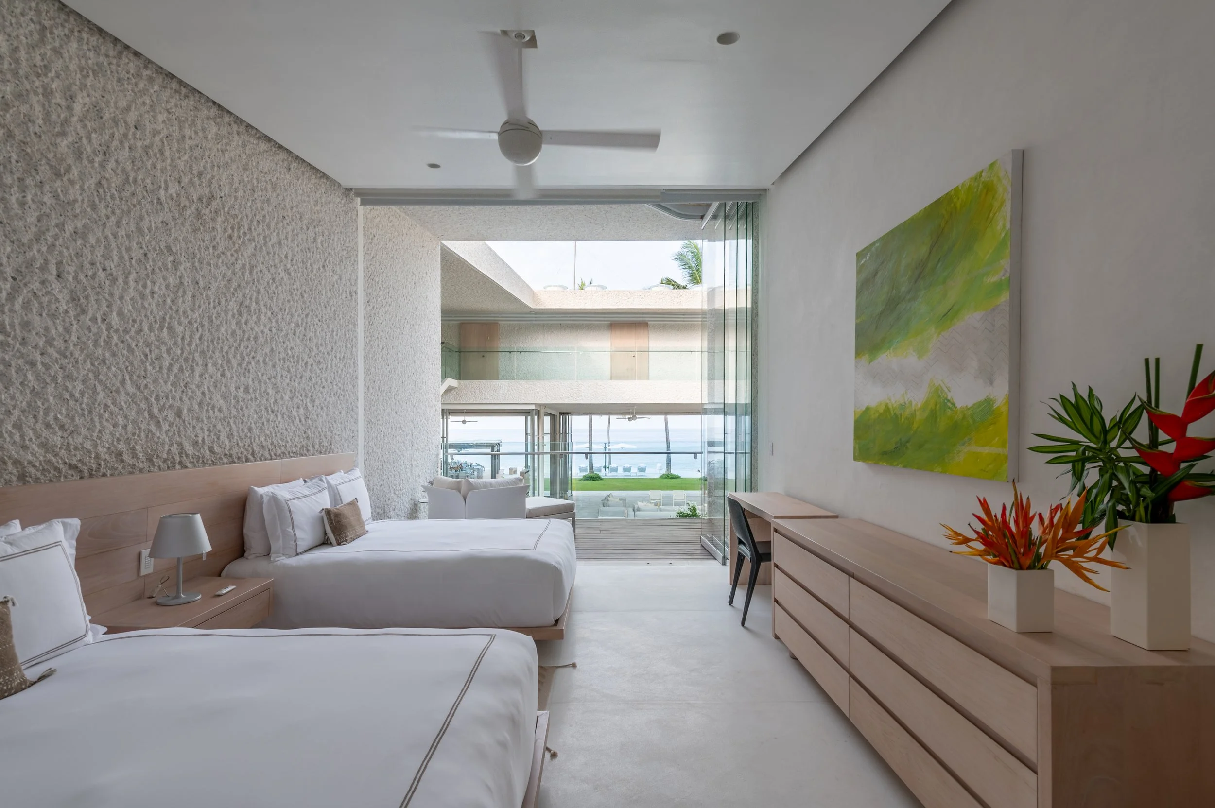 A modern, airy bedroom with two white beds, a wooden dresser with a colorful flower arrangement, a painting on the wall, and large windows overlooking a balcony with outdoor seating and ocean view.
