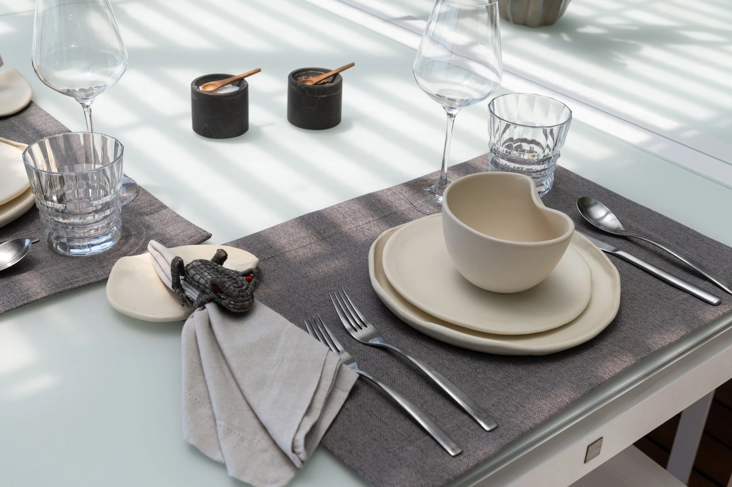 A table setting with clear water glasses, black salt and pepper shakers, white bowls, silverware, and napkins on a gray table runner, with sunlight casting shadows through window blinds.