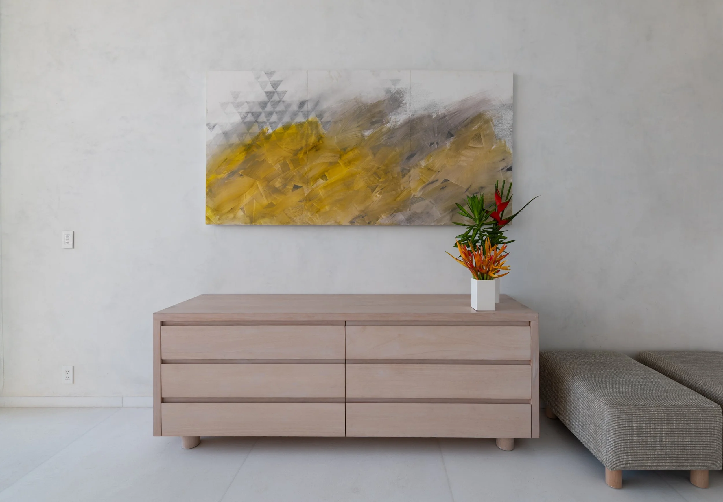 Minimalist wooden sideboard with a bouquet of tropical flowers in a white vase, above a gray sofa, and a large abstract painting with yellow, gray, and white brushstrokes hanging on a white wall.
