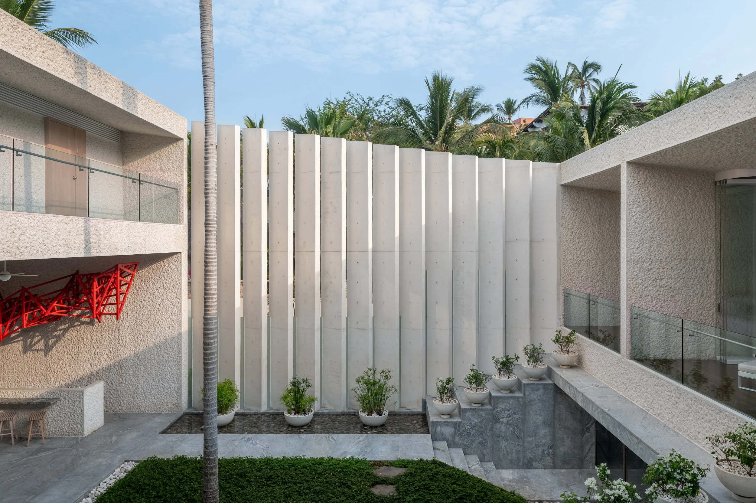 Modern residential courtyard with potted plants, marble flooring, concrete stairs, and a high white privacy wall, set against a backdrop of palm trees and a partly cloudy sky.