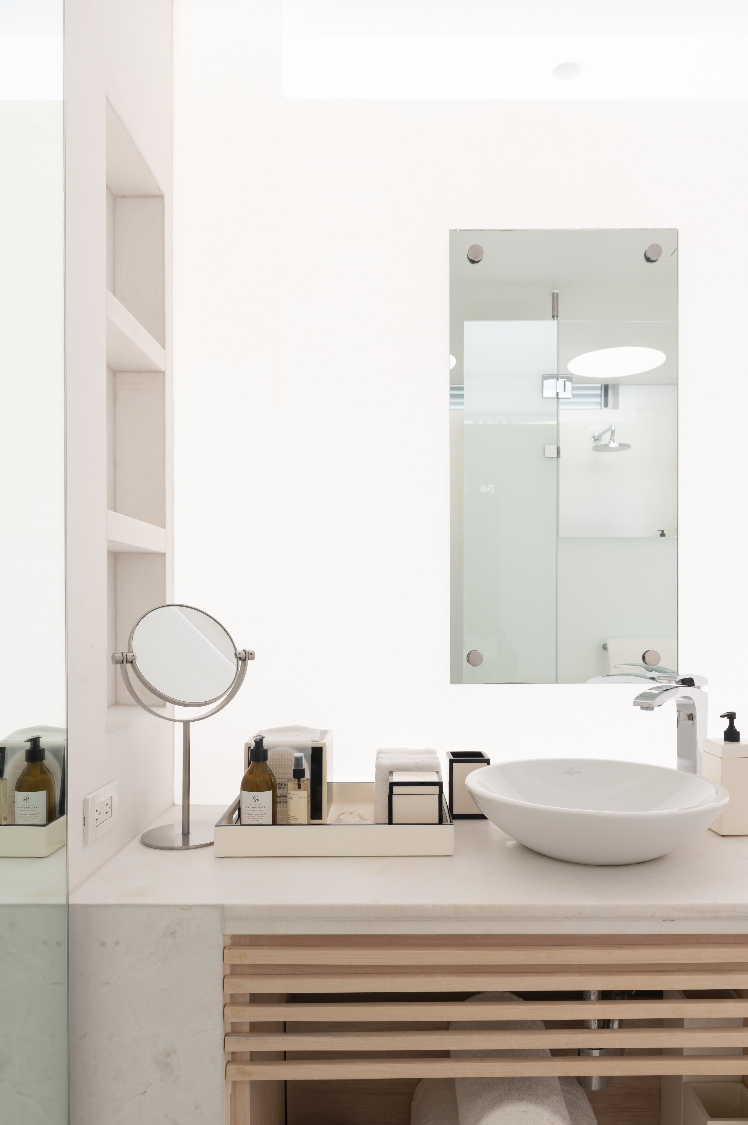 A modern bathroom with a white sink, mirror, and light-colored wooden slatted shelf underneath. Decorative bottles, towels, a mirror, and a soap dispenser are on the counter. The bathroom has white walls with built-in shelves and a glass door.
