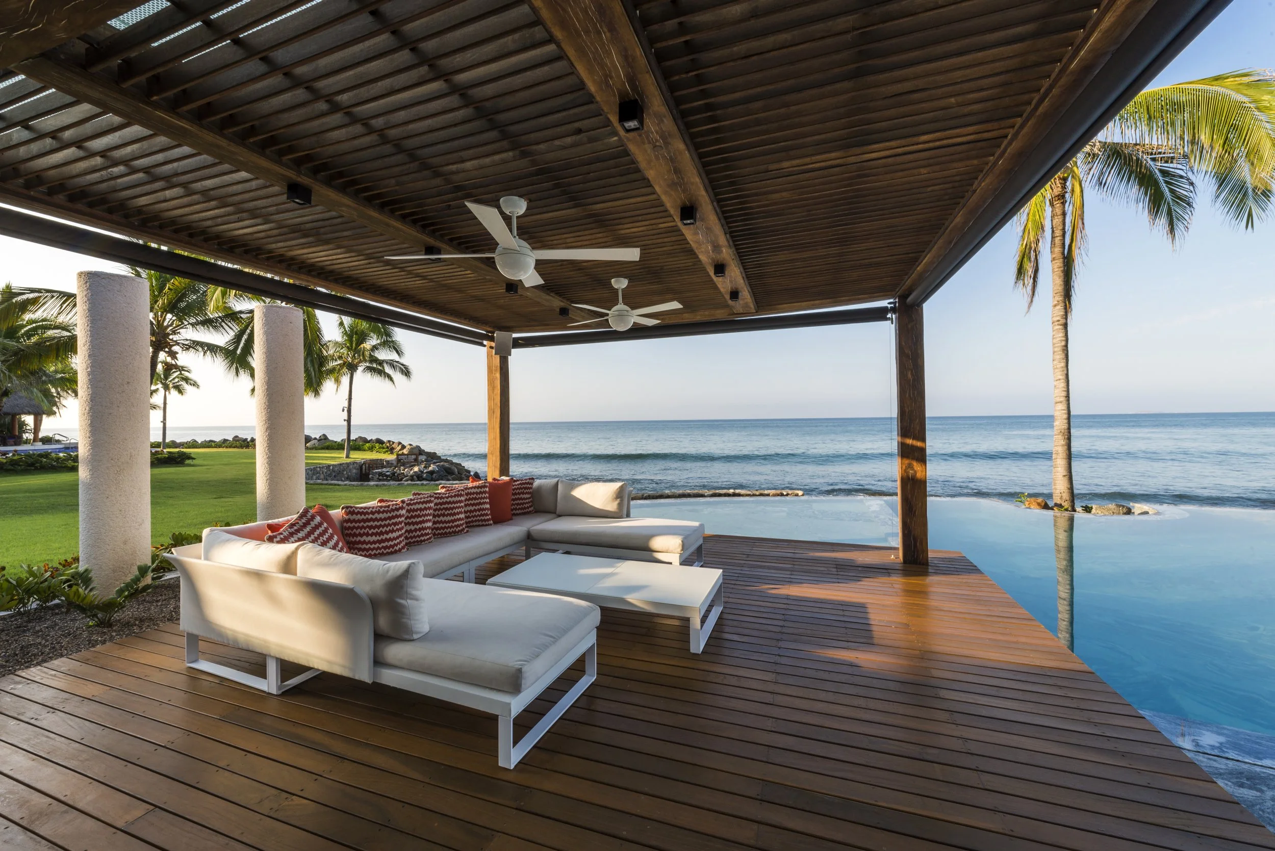 Overwater wooden deck with white outdoor furniture, ceiling fans, and view of the ocean and palm trees.
