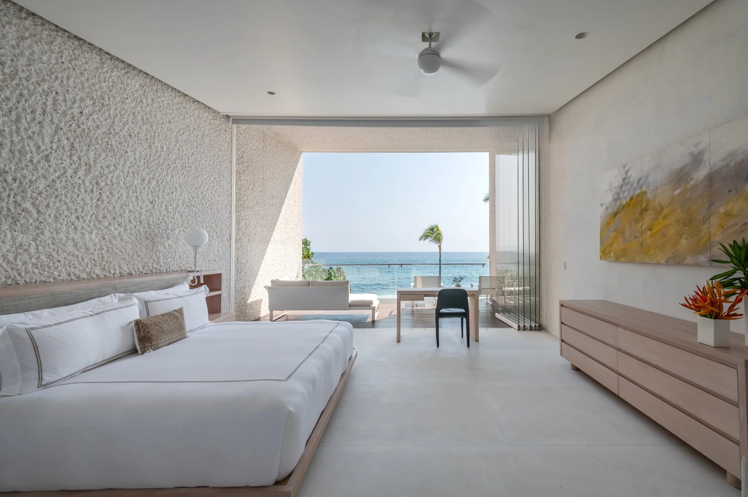 A modern bedroom with a large white bed, textured white walls, and a view of the ocean and a palm tree through an open glass wall.