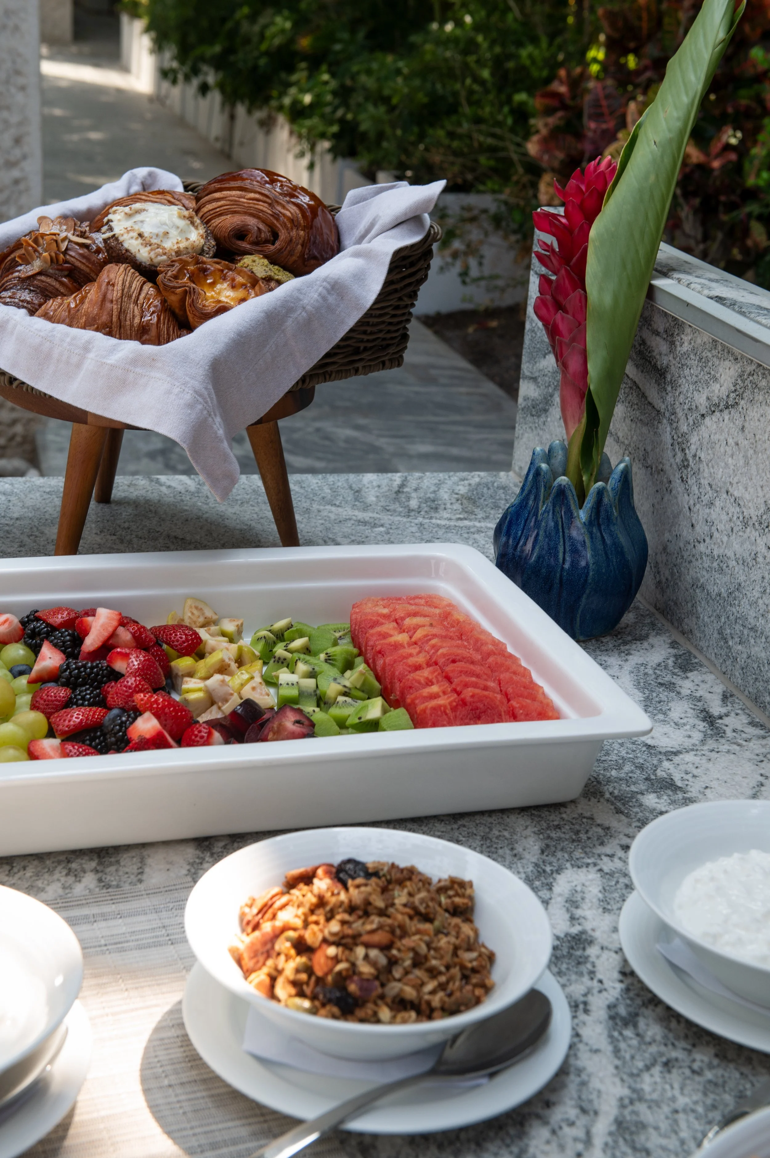 A breakfast buffet with various baked goods, fresh fruit in a white rectangular dish, and bowls of granola, yogurt, and milk on a stone countertop, with decorative red and green flowers in a blue vase in the background.