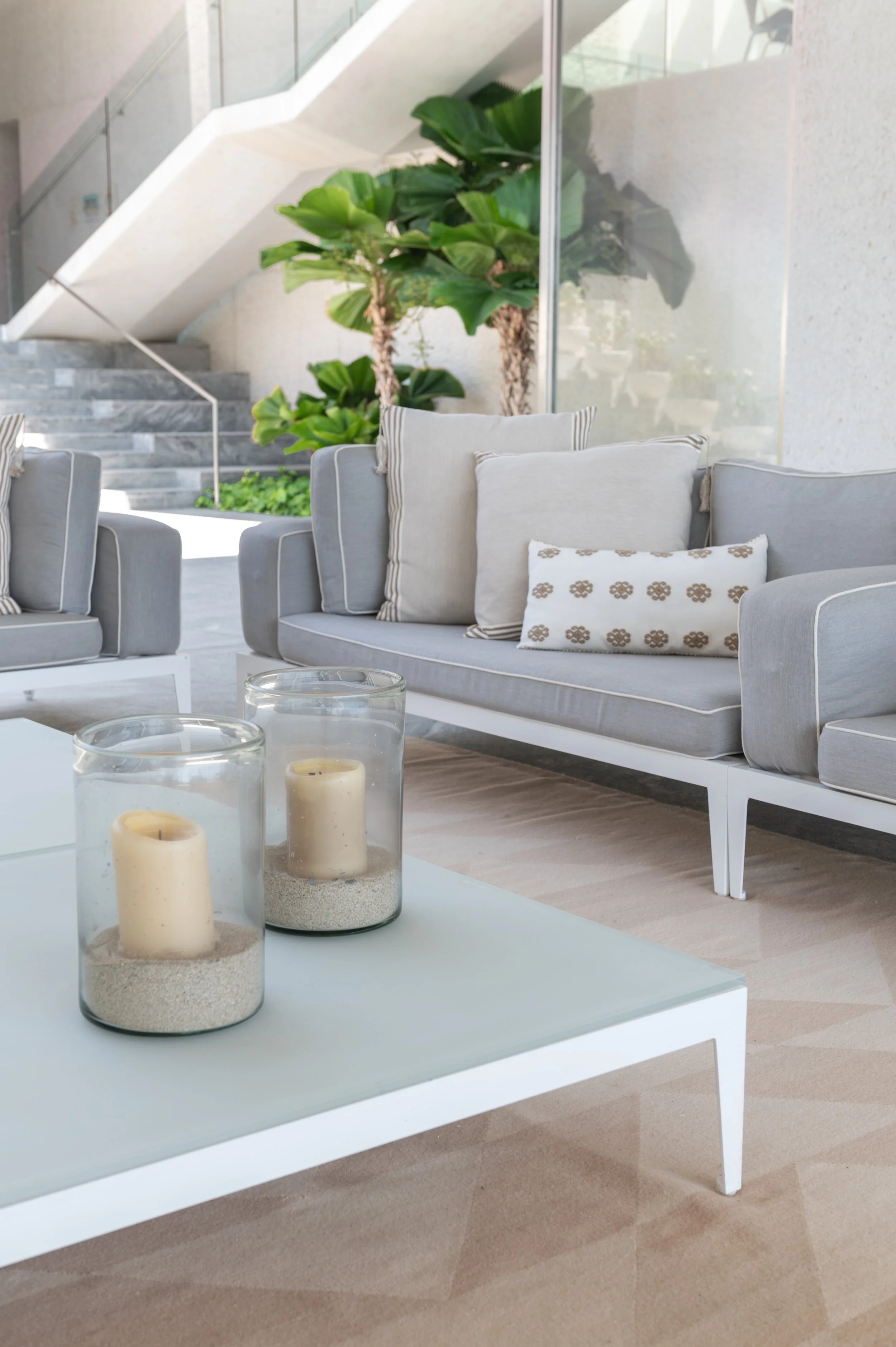 The image shows a modern, minimalist living space with a white coffee table in the foreground holding three white candles in glass holders. In the background, there are cushioned outdoor sofas with light-colored pillows, green indoor plants, and a st