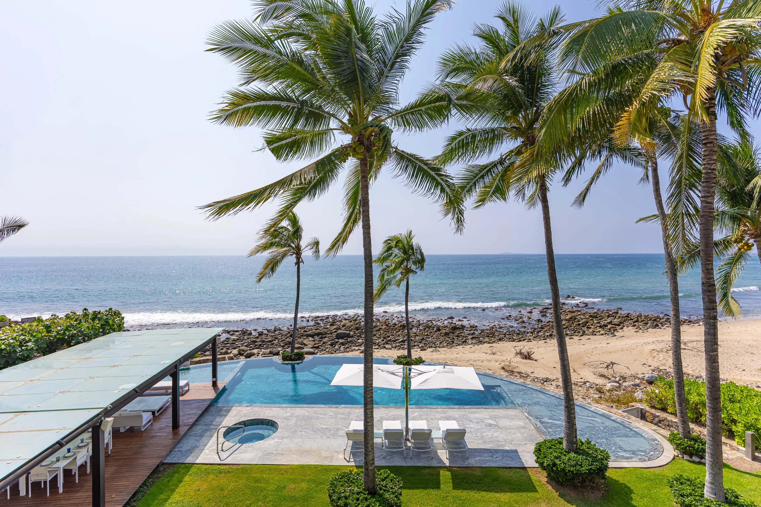 A tropical beachside scene with tall palm trees, a view of the ocean, a pool area with lounge chairs and umbrellas, and a patio with a bar and seating.