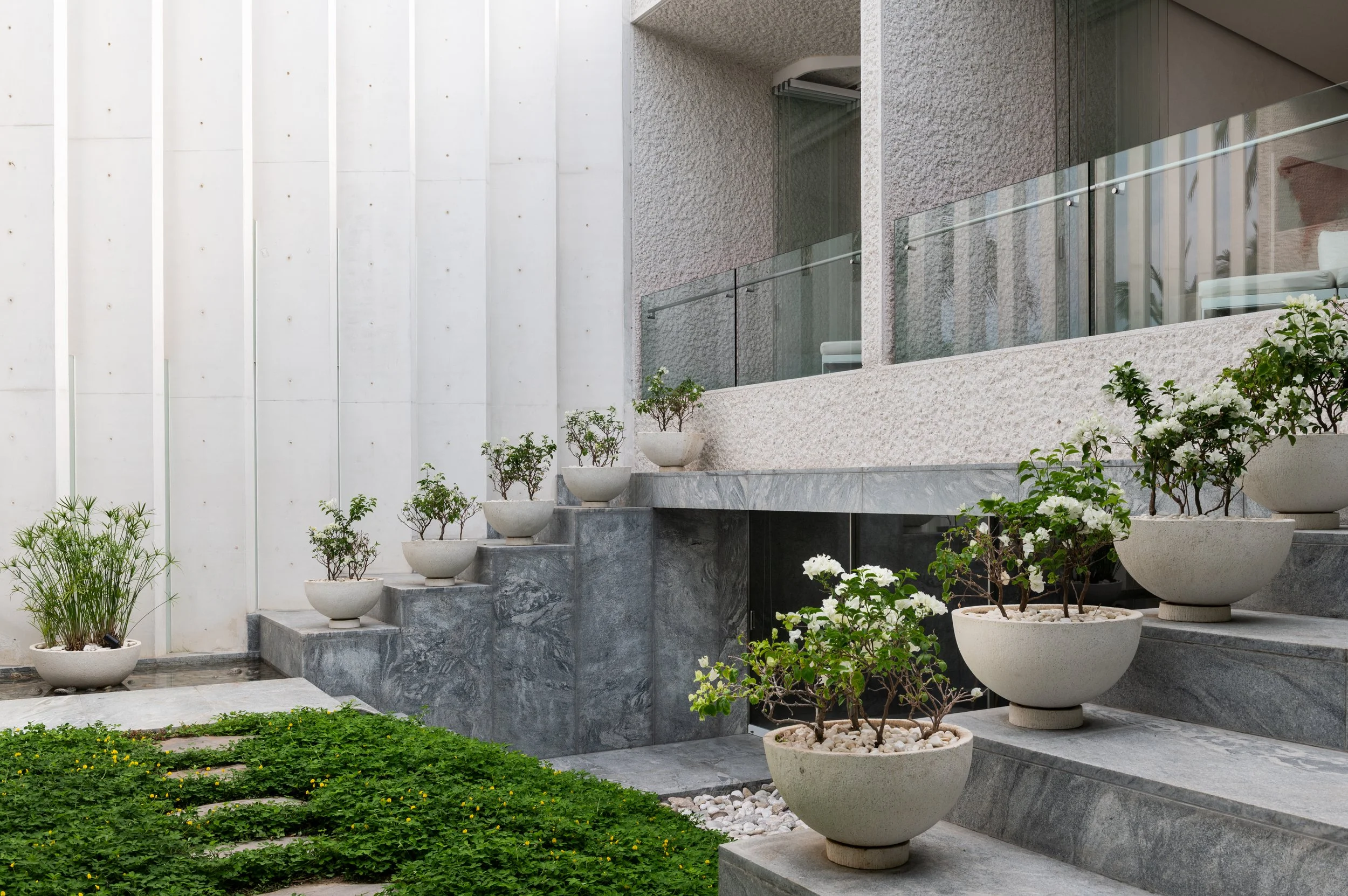 Modern house exterior with concrete stairs lined with potted plants and greenery, glass balcony, and textured wall.