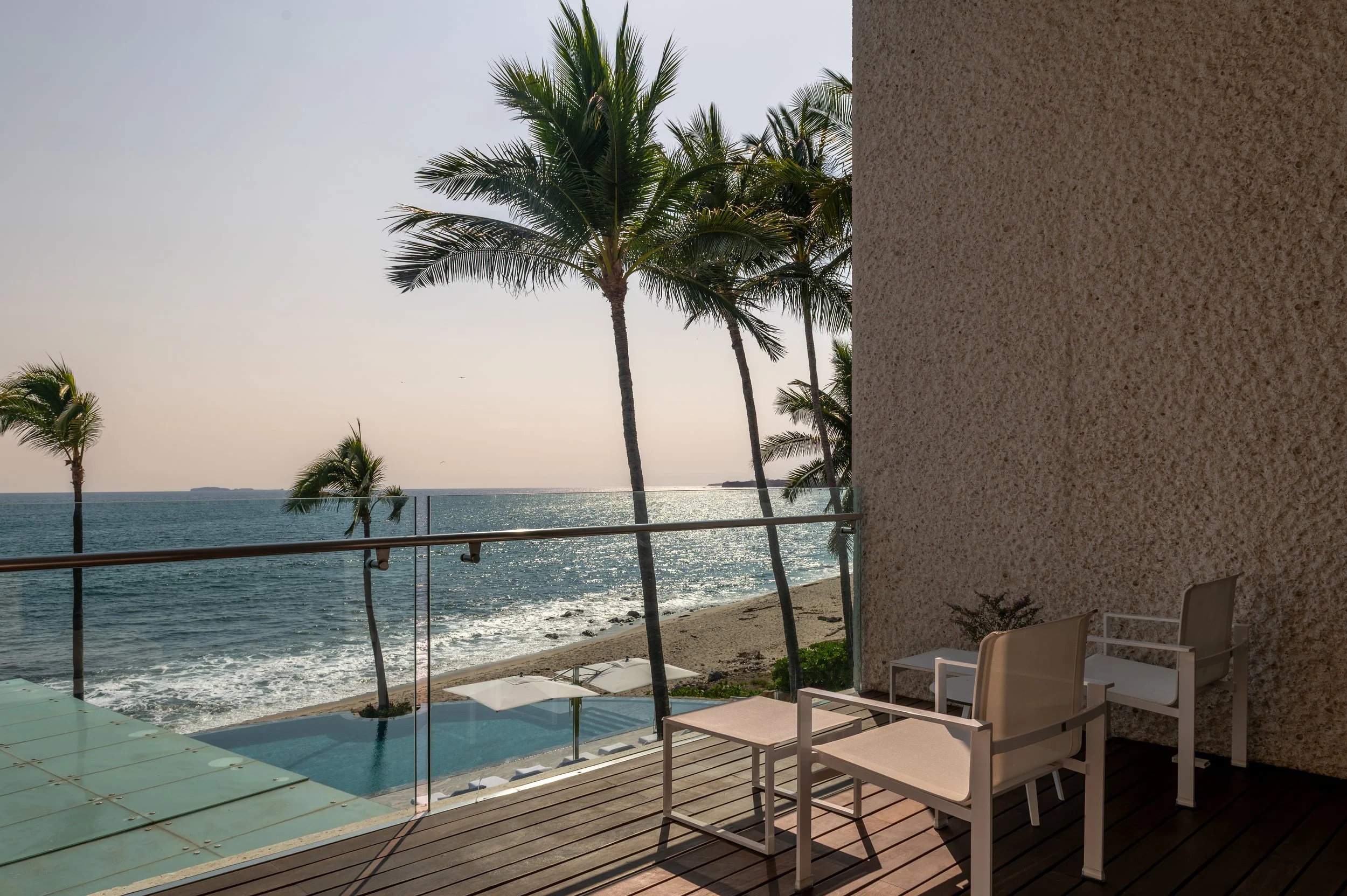 Balcony view overlooking the ocean with palm trees and a pool in the foreground, with outdoor chairs and a glass railing