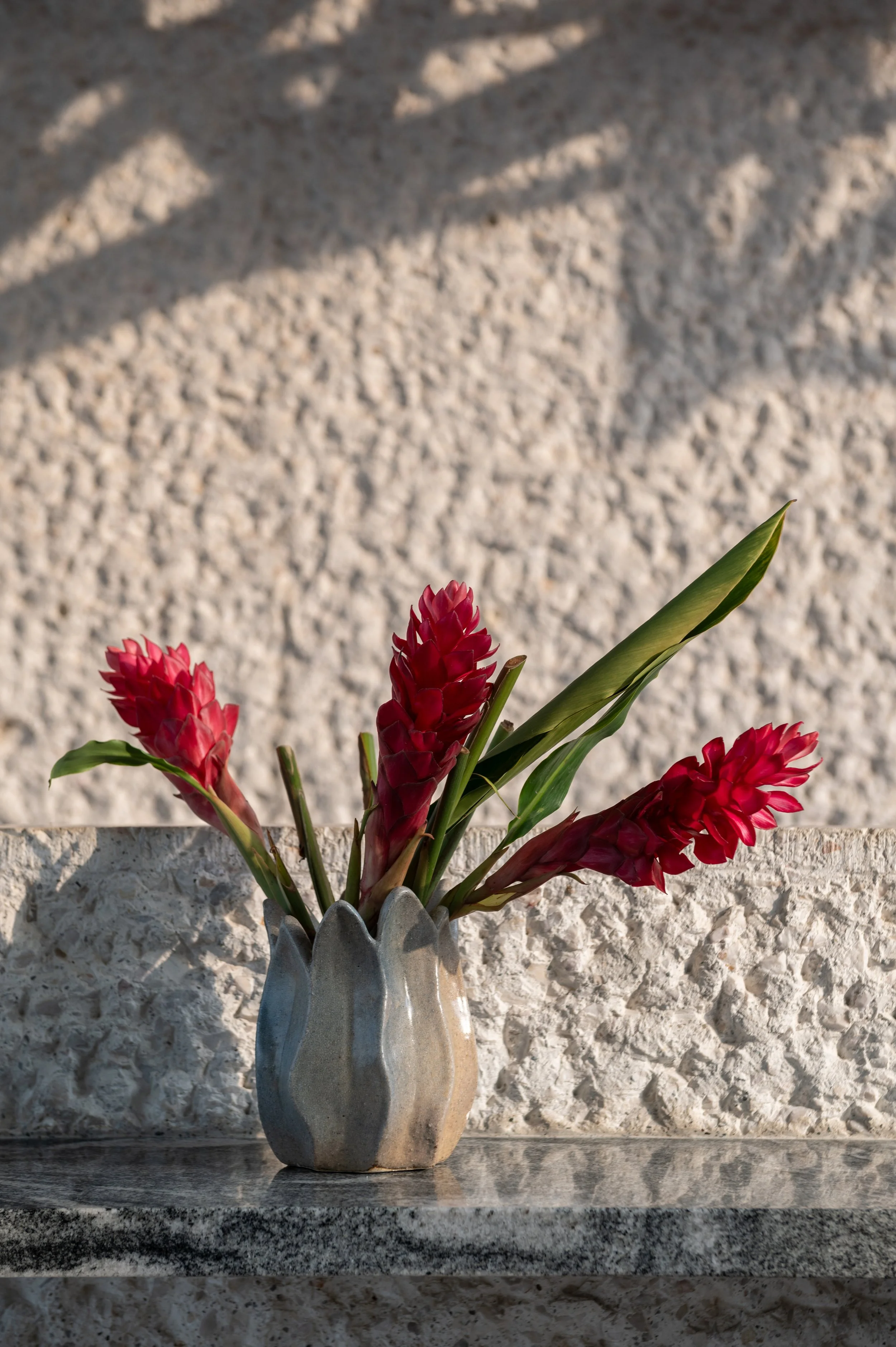 A ceramic vase with a ribbed, petal-like design holds pink ginger flowers and green leaves, placed on a dark granite surface against a textured beige stone wall.