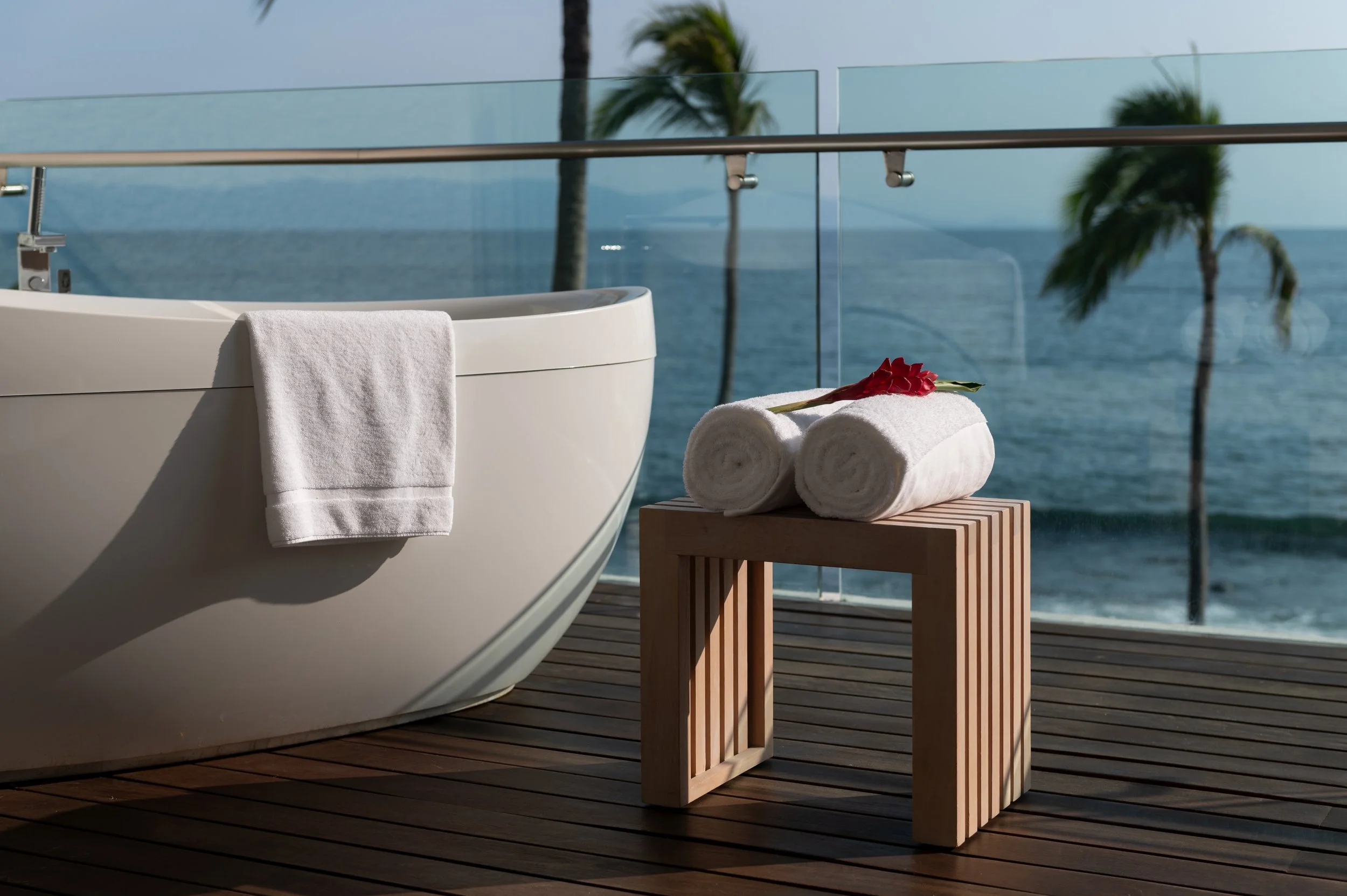A white bathtub with a towel draped over its side, a rolled towel with a red flower on top on a wooden stool, set on a wooden deck with ocean and palm trees in the background.