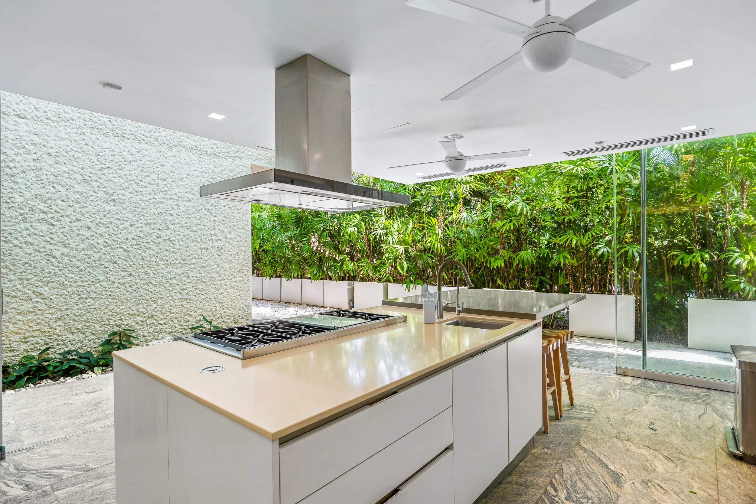 Modern kitchen with white cabinets, a large beige countertop, a stainless steel stove, and a vent hood, with a large window showing lush green plants outside.