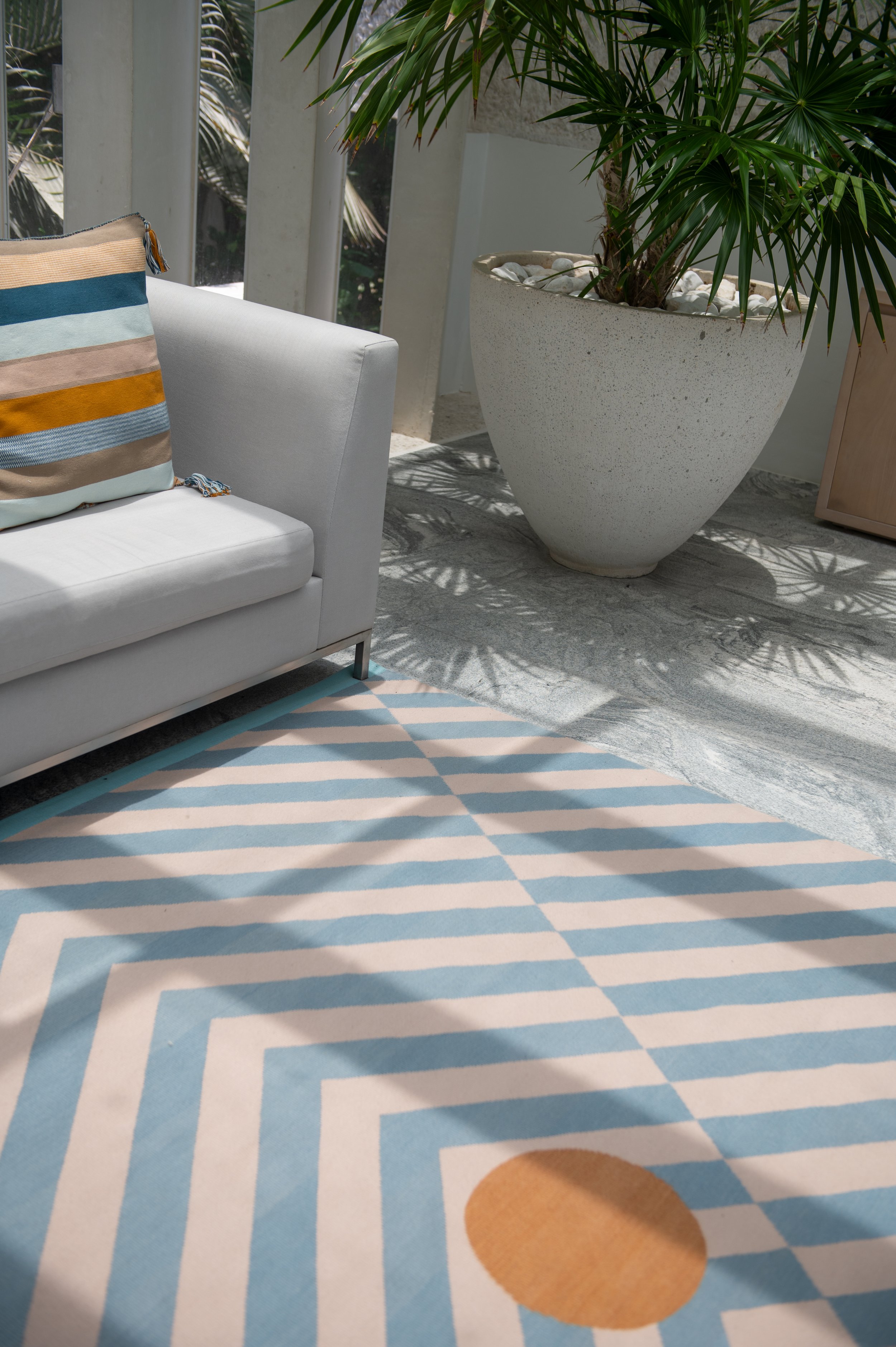 Close-up of a patterned carpet with blue and white stripes and a large orange circle, next to white sofa with multicolored striped cushion, and tall potted green plant with white stones in a large beige pot.