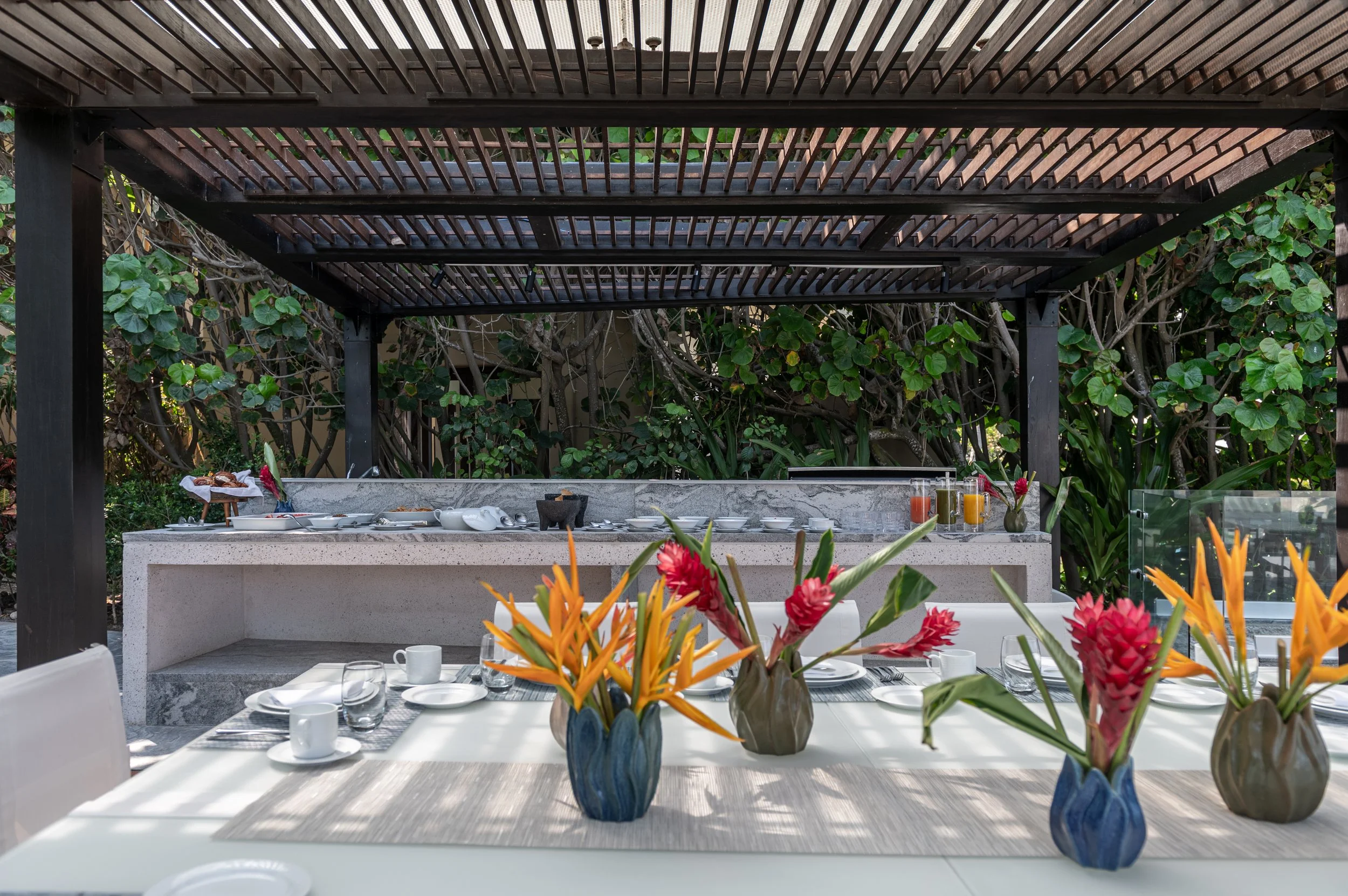 Outdoor dining area with a table set with white plates, glasses, and cups, decorated with a floral centerpiece featuring orange and pink flowers, beneath a dark wooden pergola with greenery in the background.