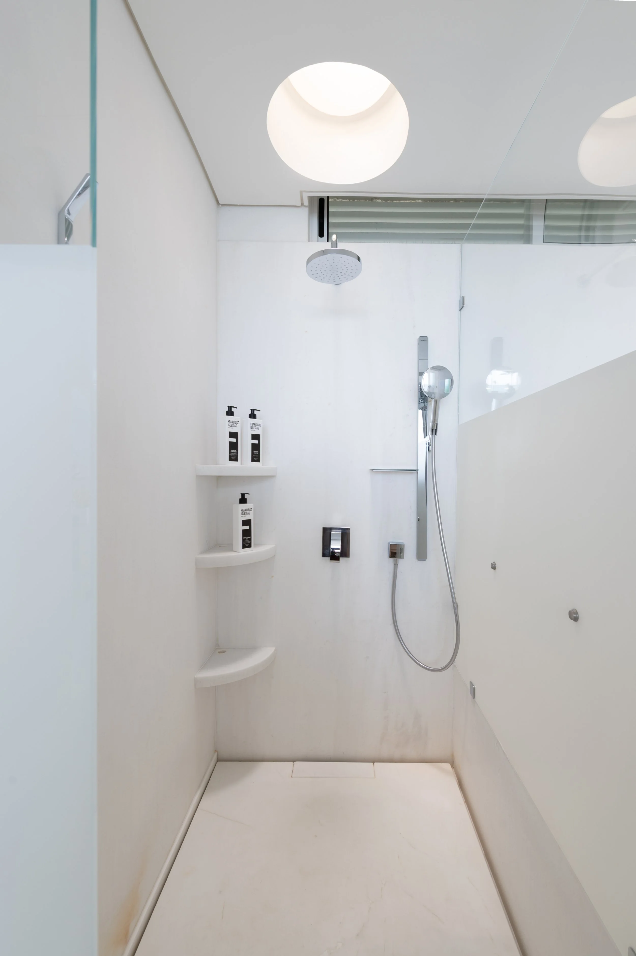 Modern minimalist shower with white walls and floor, rain showerhead, handheld shower, built-in corner shelves with shampoo bottles, and clean design.
