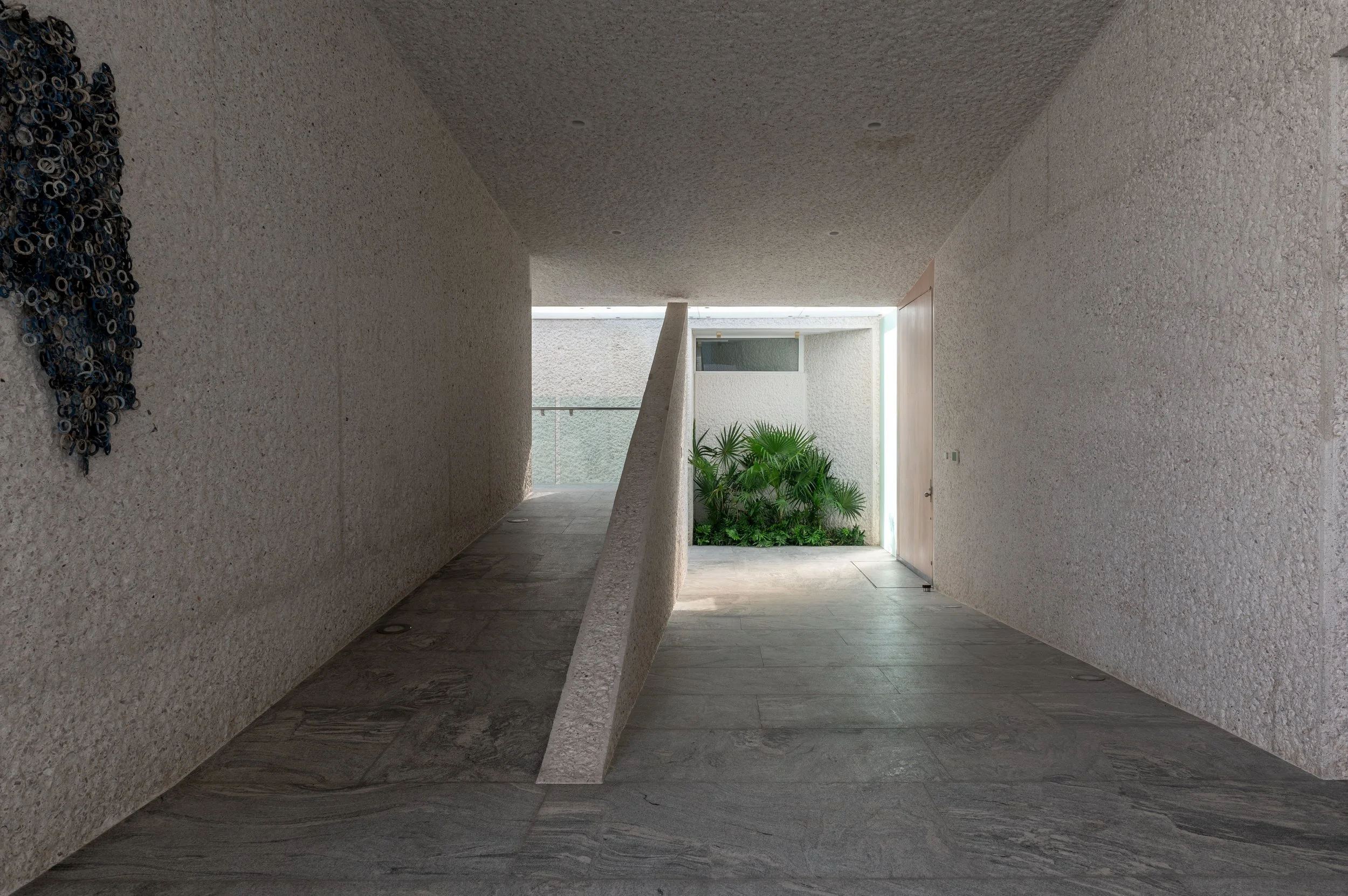 A modern, minimalist interior space with textured off-white walls and ceiling, a concrete railing, a potted green plant, and a door leading outside with natural light.