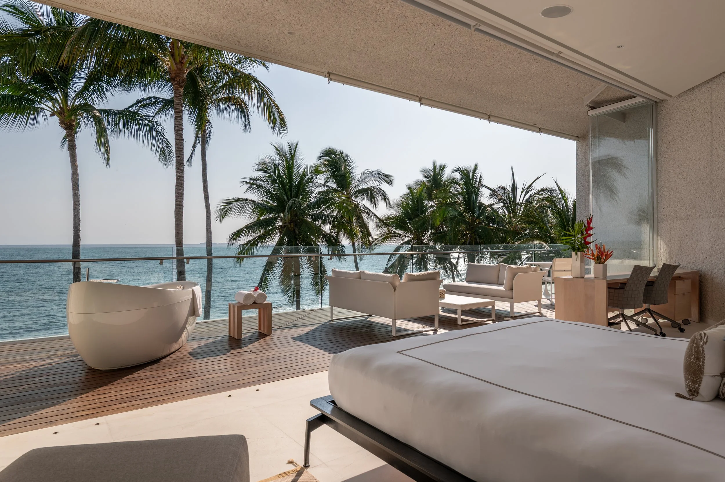 Modern indoor lounge with white sofas and chairs, overlooking a balcony with palm trees and an ocean view.