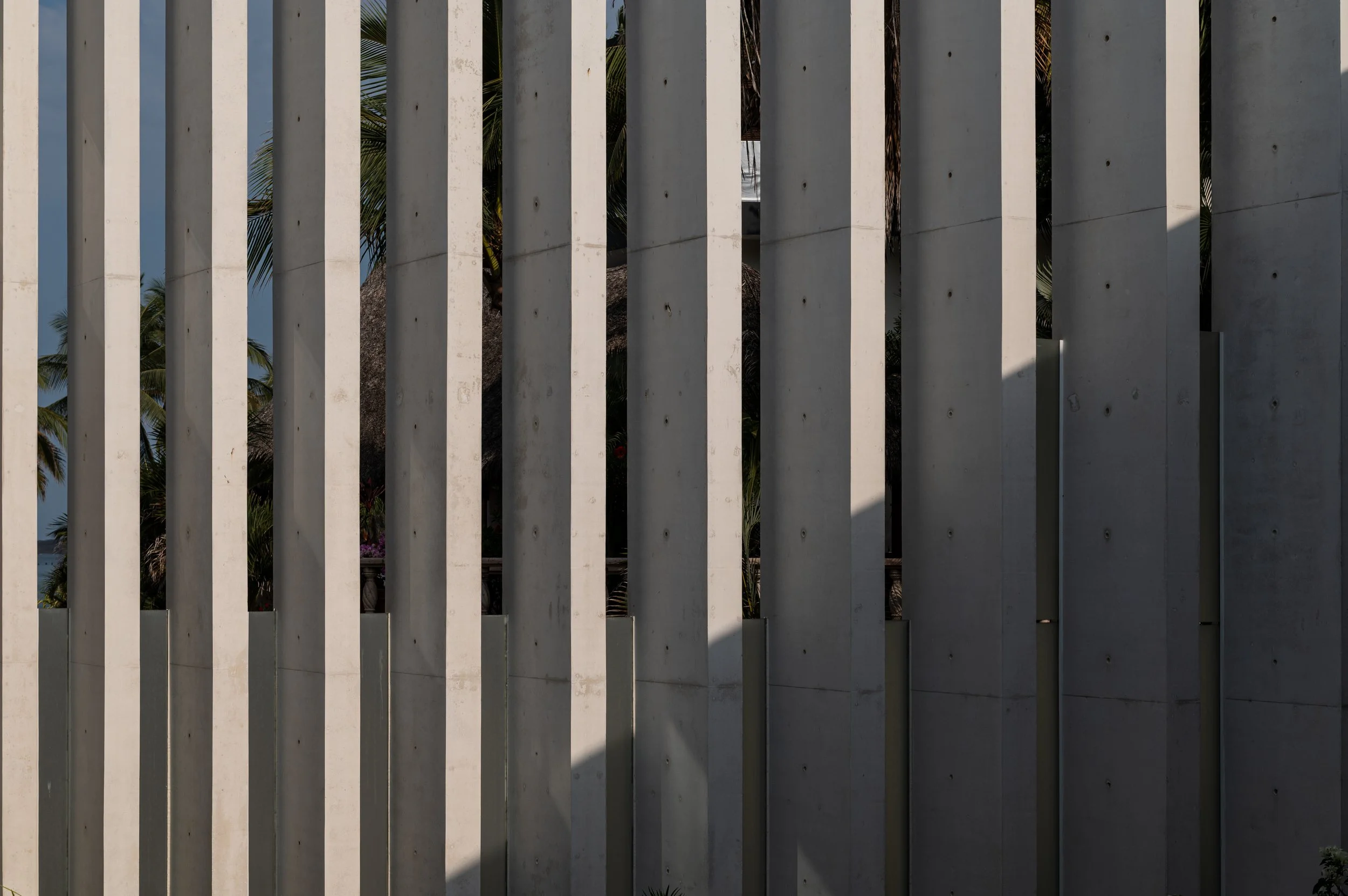 Vertical concrete slats creating a fence with glimpses of palm trees and a thatched roof building behind, against a blue sky.