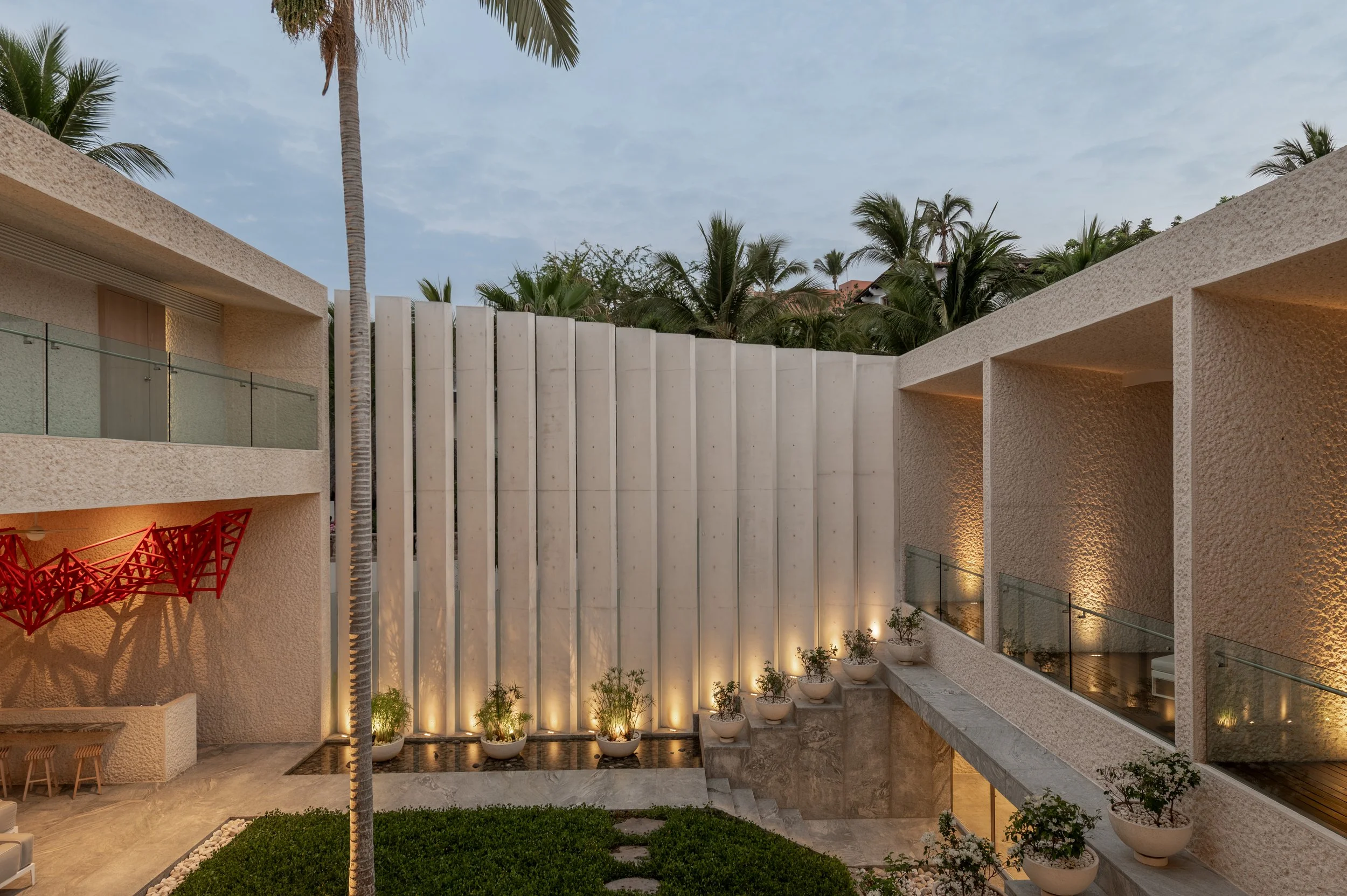 Modern building with textured beige walls, glass railings, and tall palm trees, illuminated by outdoor lighting at dusk.