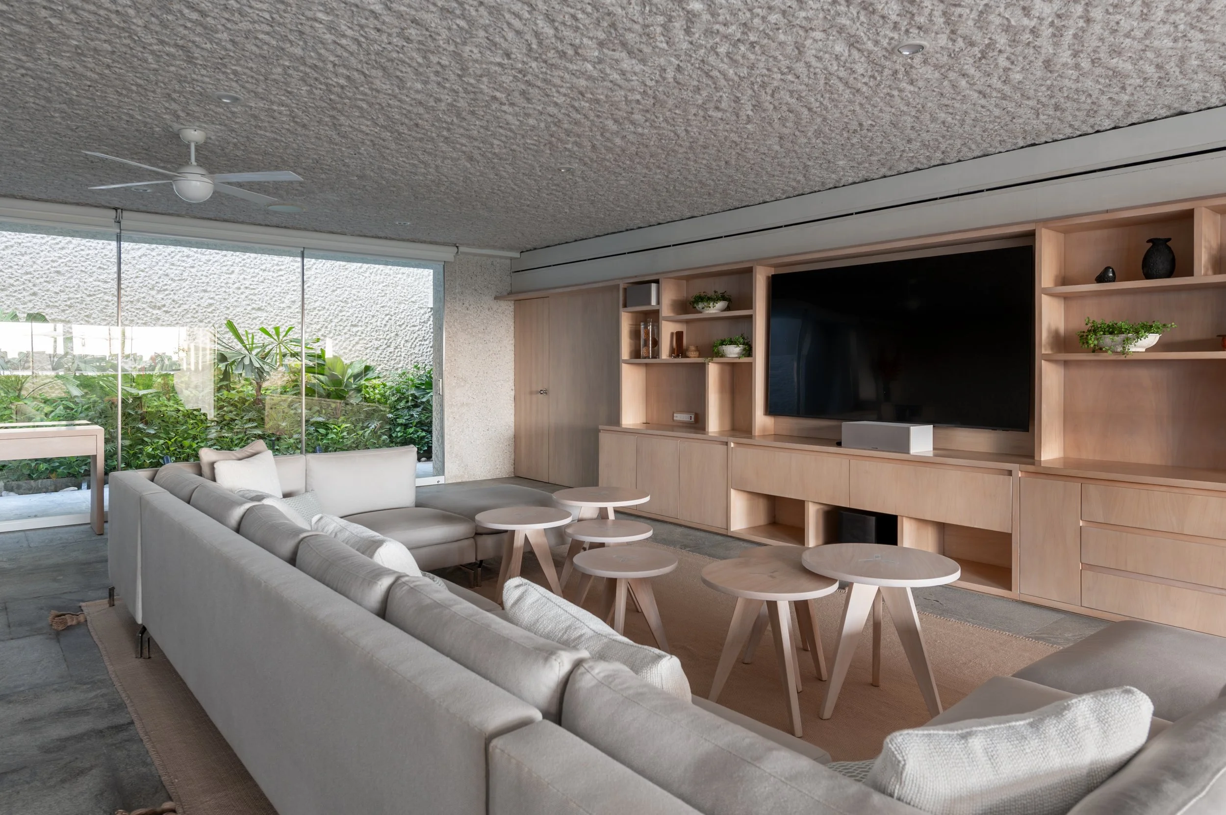 Living room with large sliding glass doors leading to greenery outside, white sofas, round wooden tables, built-in wooden shelving with decorative items, and a ceiling fan.